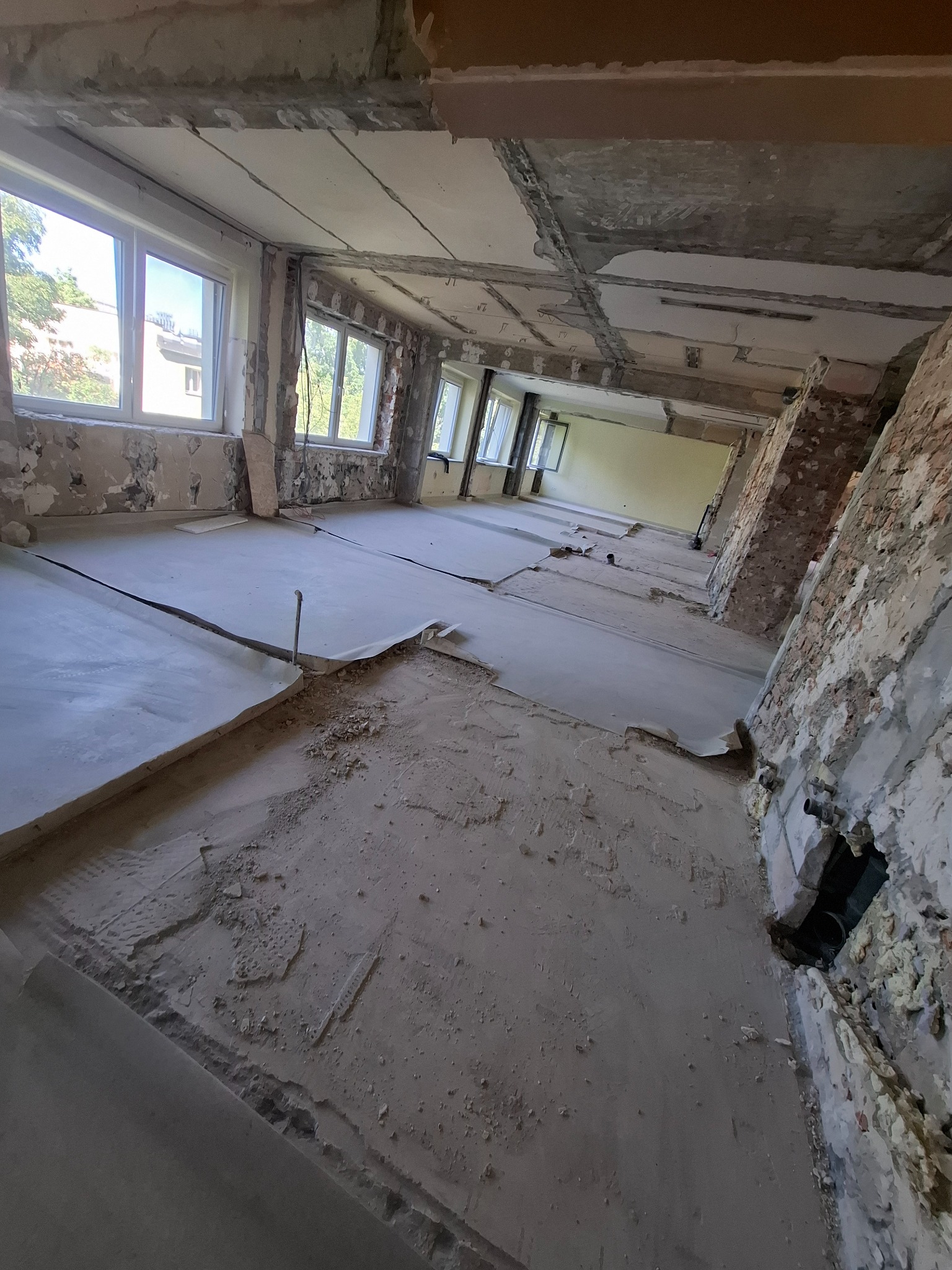 Wnętrze w trakcie rozbiórki. Widoczne odsłonięte cegły, fragmenty instalacji, okna z widokiem na zieleń i resztki podłogi. Prace rozbiórkowe w pomieszczeniu.