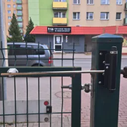 Zielona furtka z automatycznym zamkiem na tle miejskiego krajobrazu z budynkami i zaparkowanym vanem. Widoczny mechanizm zamykający, chodnik z kostki.