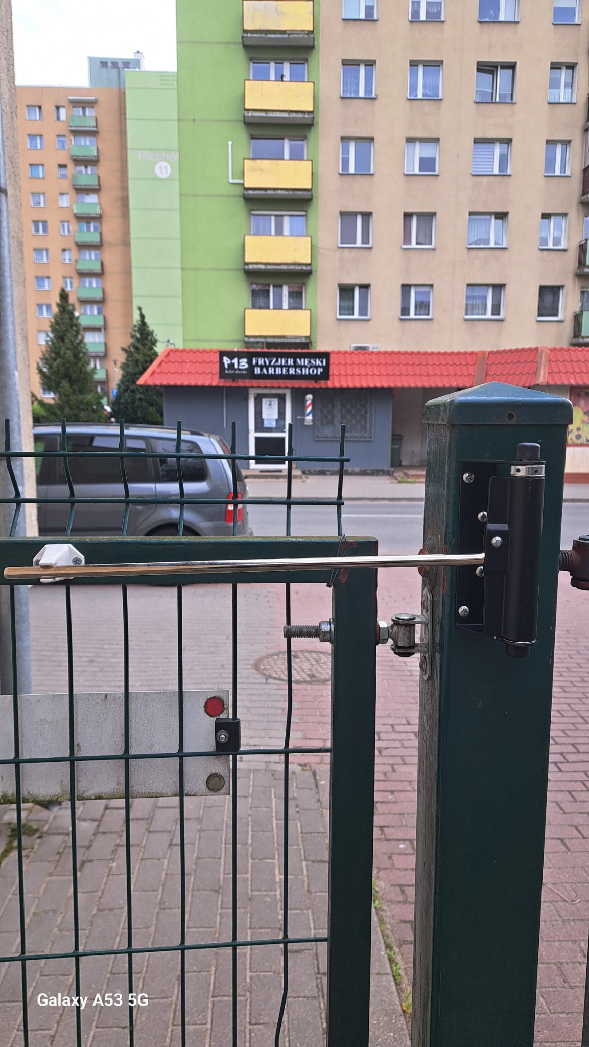 Zielona furtka z automatycznym zamkiem na tle miejskiego krajobrazu z budynkami i zaparkowanym vanem. Widoczny mechanizm zamykający, chodnik z kostki.