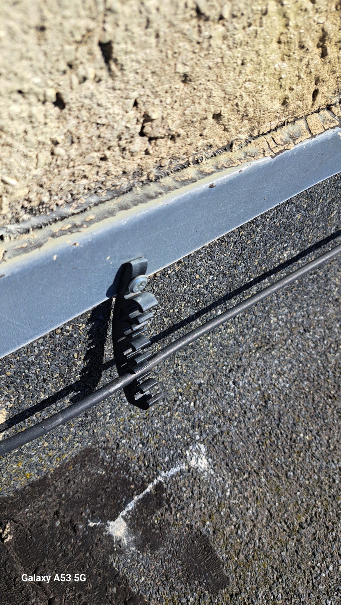 Szary przewód przymocowany do ściany za pomocą plastikowego uchwytu z metalową śrubą. Widoczna struktura ściany i pokrycie dachu z drobnym kruszywem.