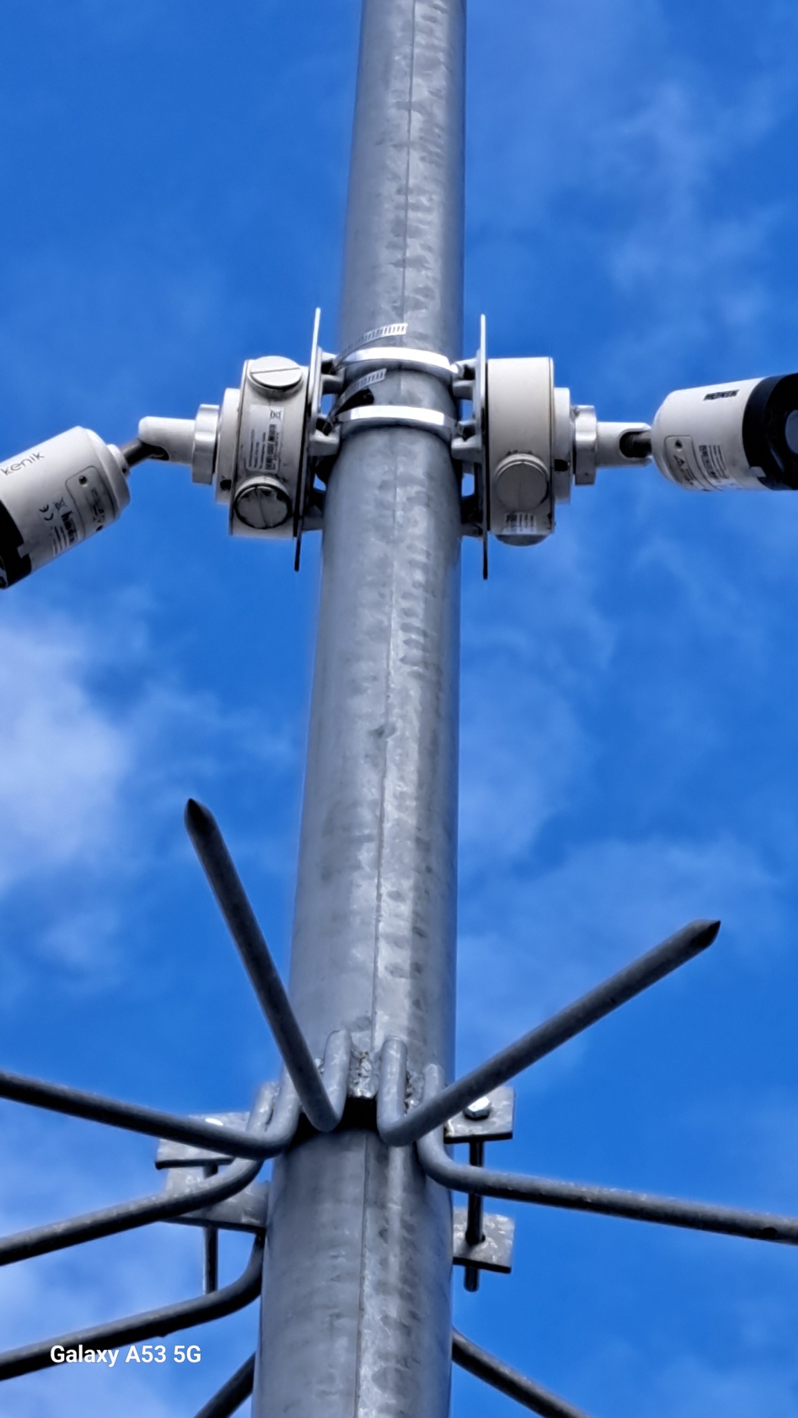 Słup z zamontowanymi kamerami przemysłowymi marki Kenik i zabezpieczeniem anty-ptasim na tle błękitnego nieba. Widok z dołu.