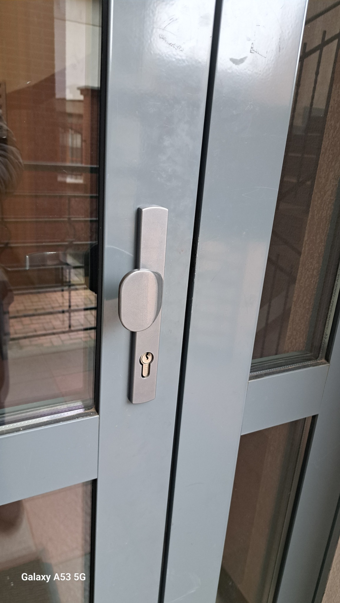 Szare drzwi wejściowe z szybą i srebrną klamką z zamkiem, widoczne z bliska, zarys wnętrza budynku w odbiciu szyby.