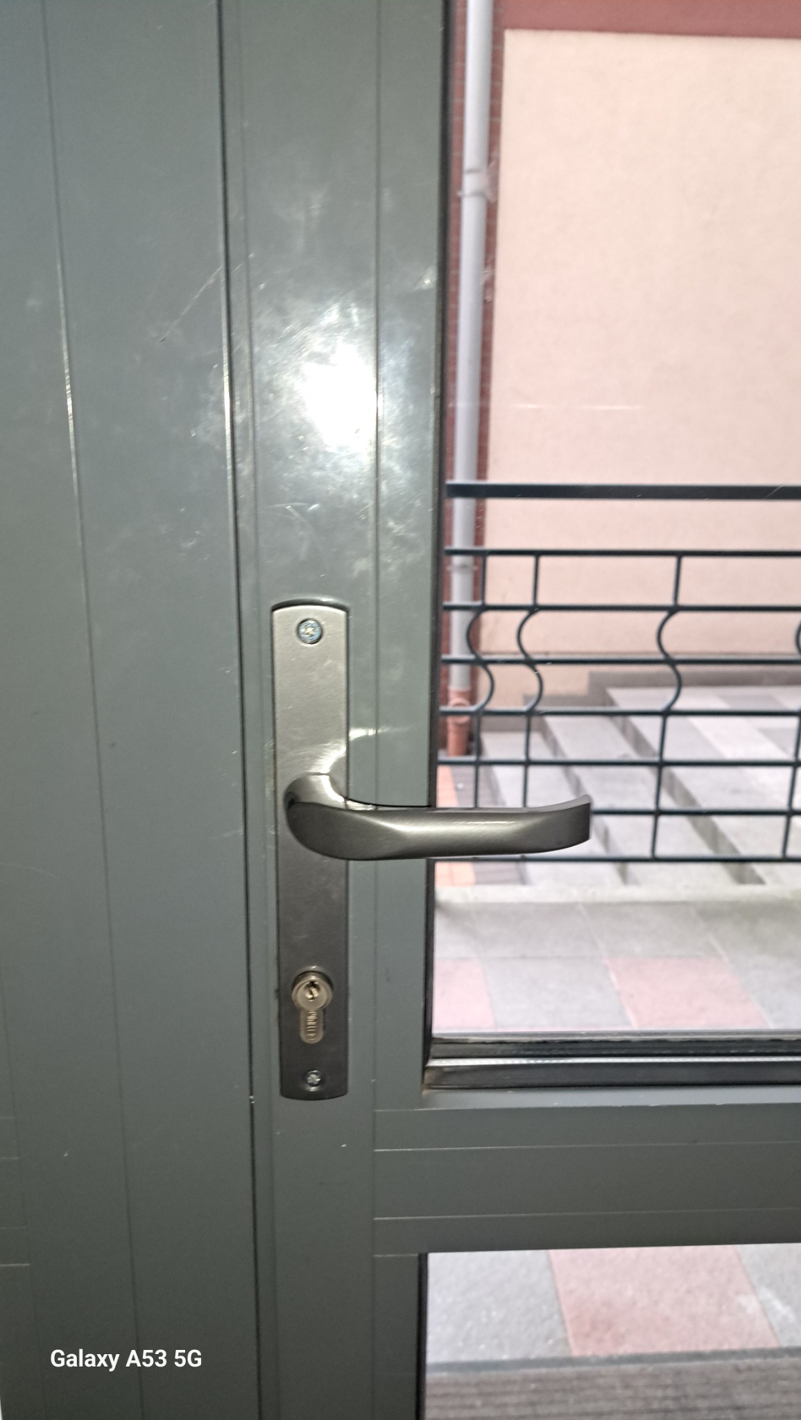 Szare drzwi balkonowe z metalową klamką i zamkiem, widok na balkon z ozdobną kratą. Detal klamki, zarysowania na drzwiach.