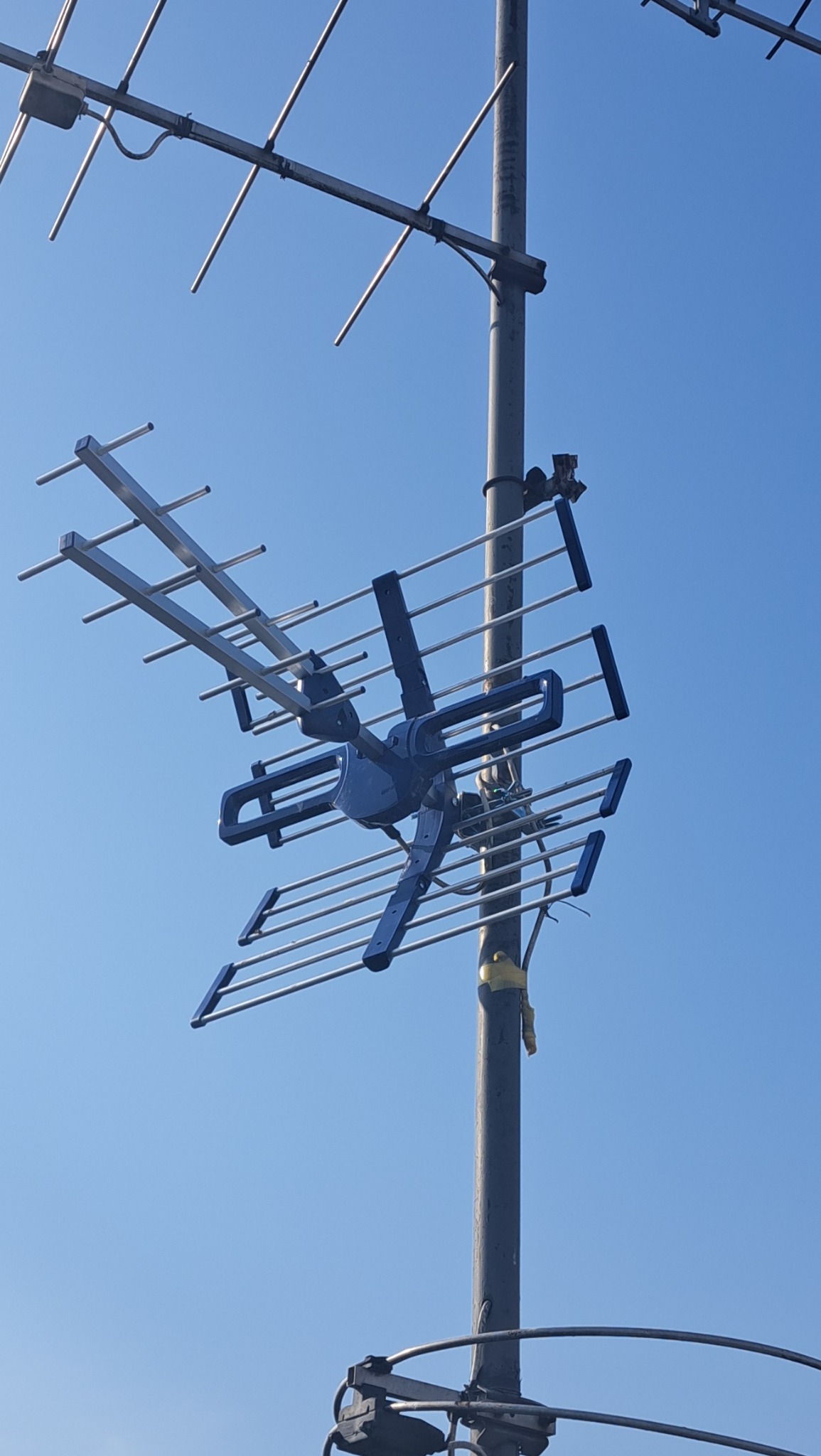 Antena telewizyjna na maszcie, skierowana ku błękitnemu niebu. Widoczne elementy konstrukcyjne i kable. Detaliczny widok na system odbioru sygnału.