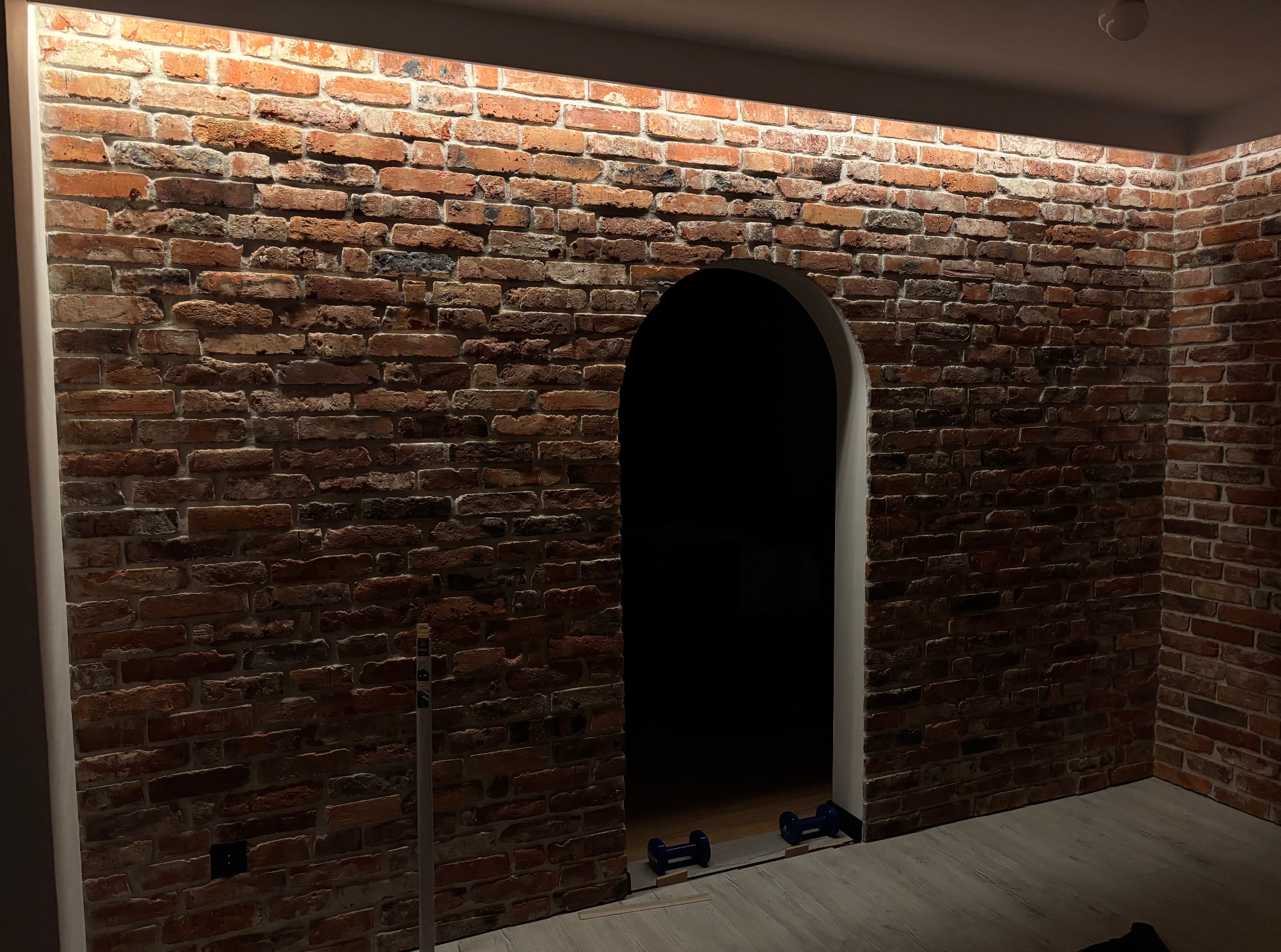 Ściana z cegły dekoracyjnej z łukowatym przejściem, oświetlona od góry. Widoczne detale faktury cegieł i subtelne oświetlenie. W przejściu widoczne hantle.