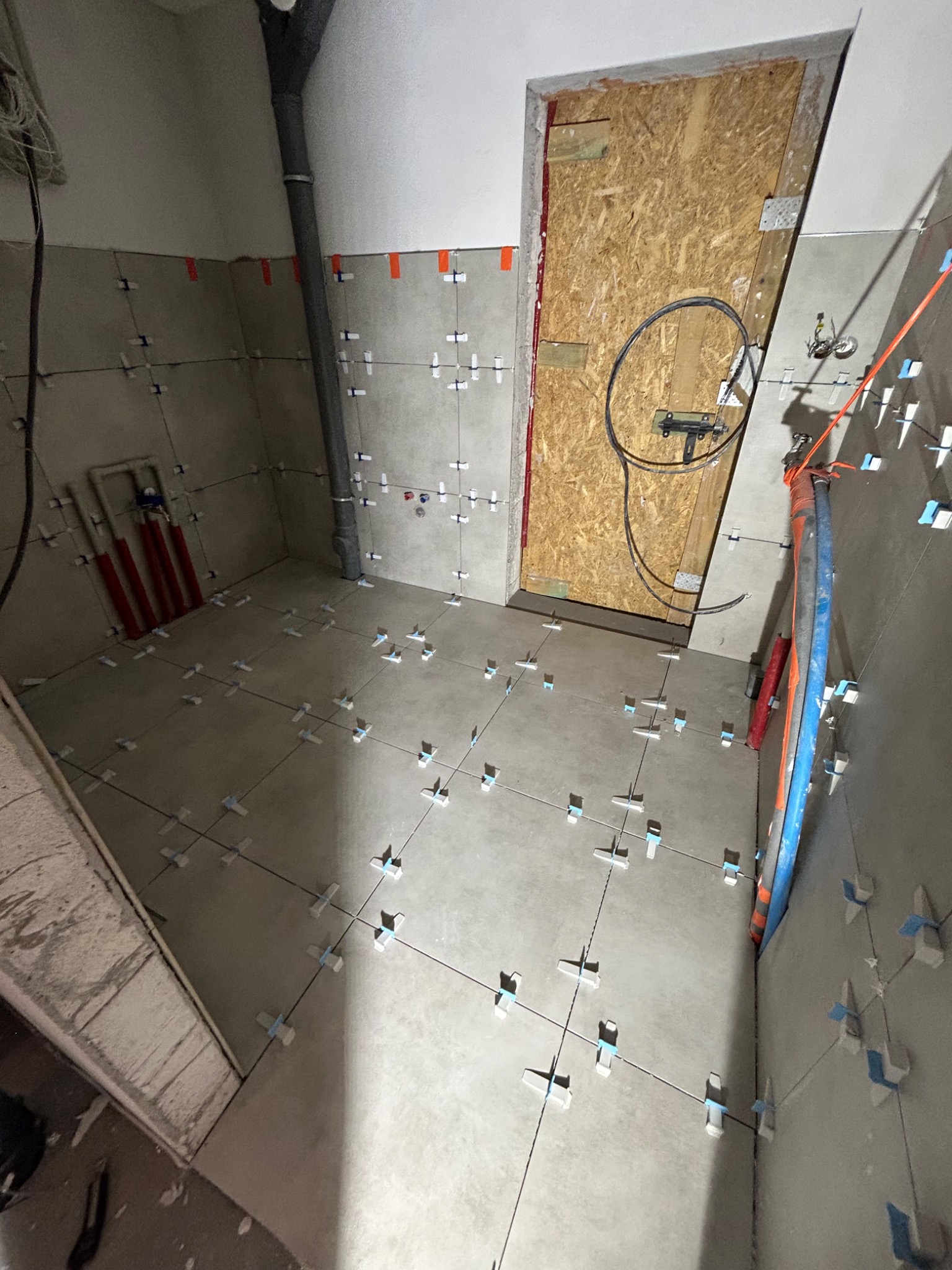 Szare płytki podłogowe i ścienne w trakcie układania. Krzyżyki dystansowe, widoczne rury i instalacje. Surowy stan wykończenia łazienki w trakcie prac glazurniczych.