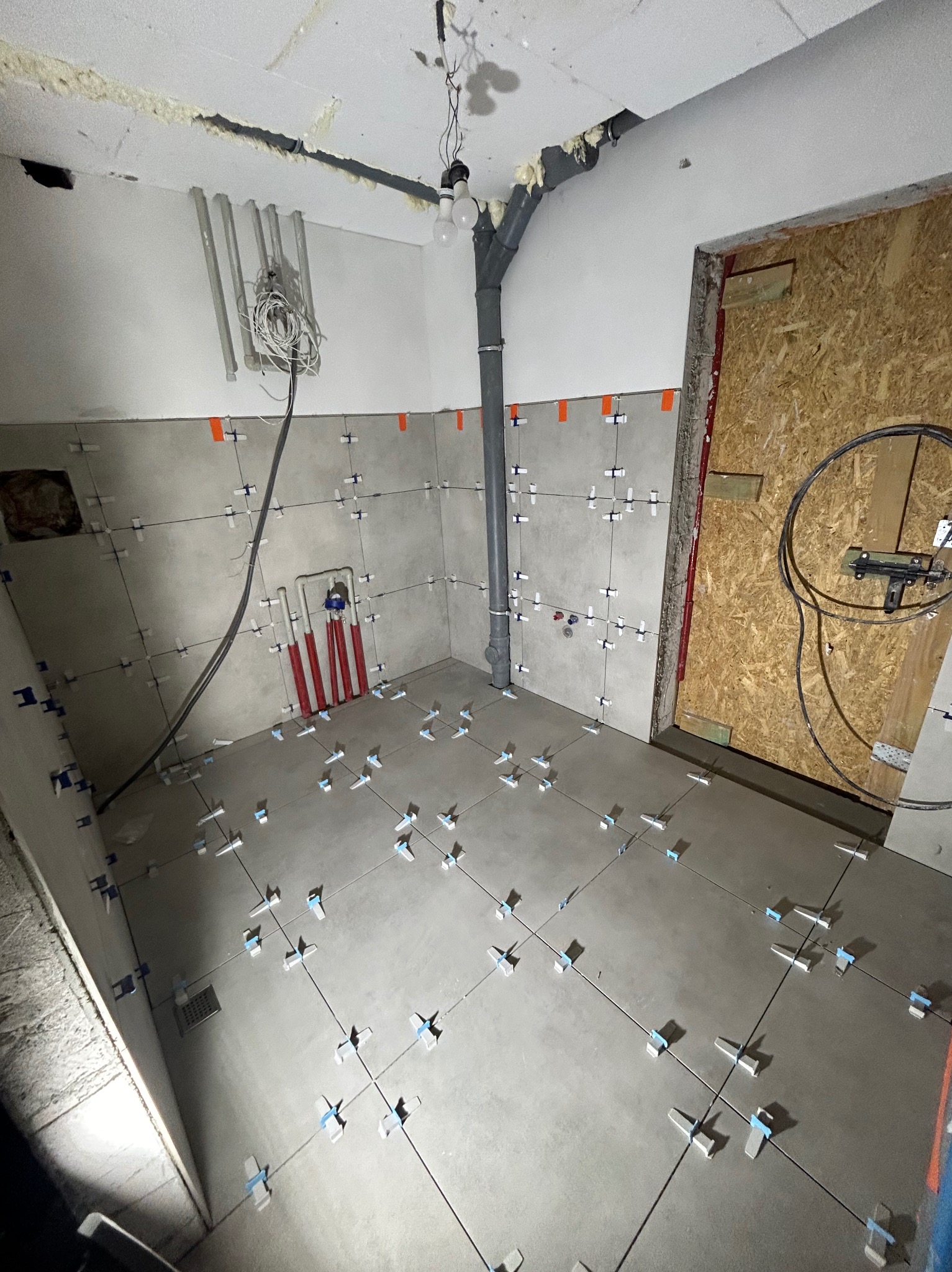 Kładzenie szarych płytek na podłodze i ścianach w łazience, z krzyżykami dystansowymi. Widoczne rury i instalacje, surowy stan wykończenia.