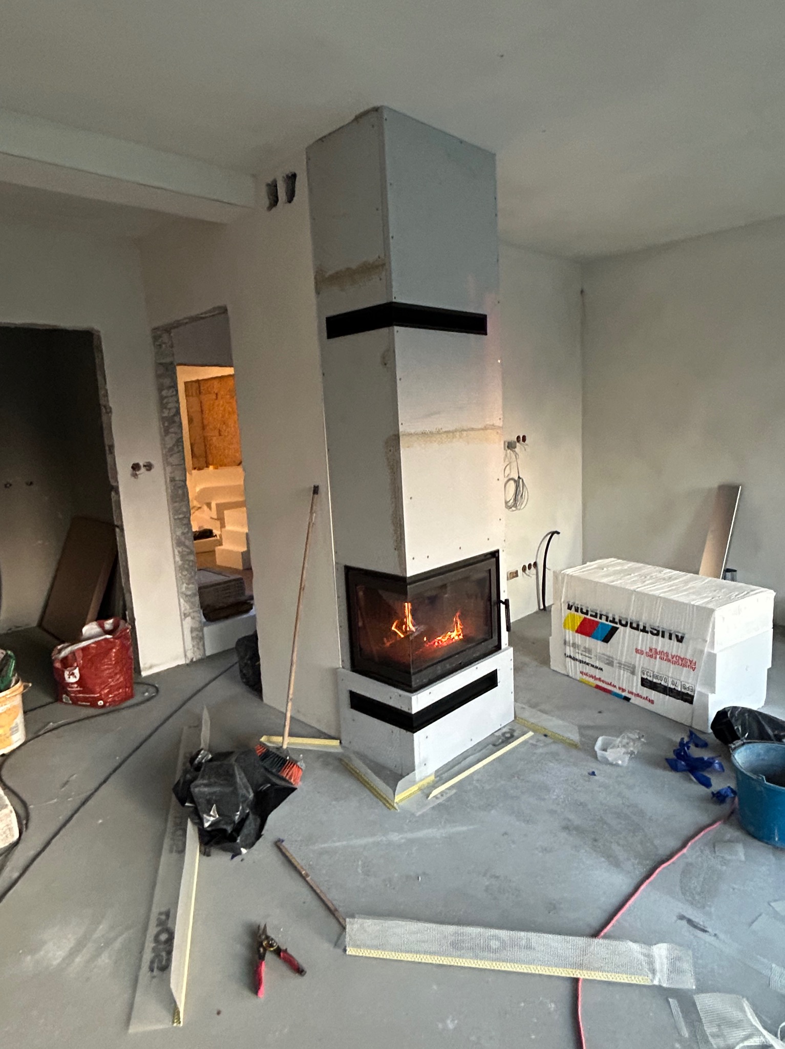 Wnętrze w trakcie prac wykończeniowych z widocznym kominkiem obudowanym płytami g-k, narzędziami i materiałami budowlanymi. Widoczne przewody elektryczne.