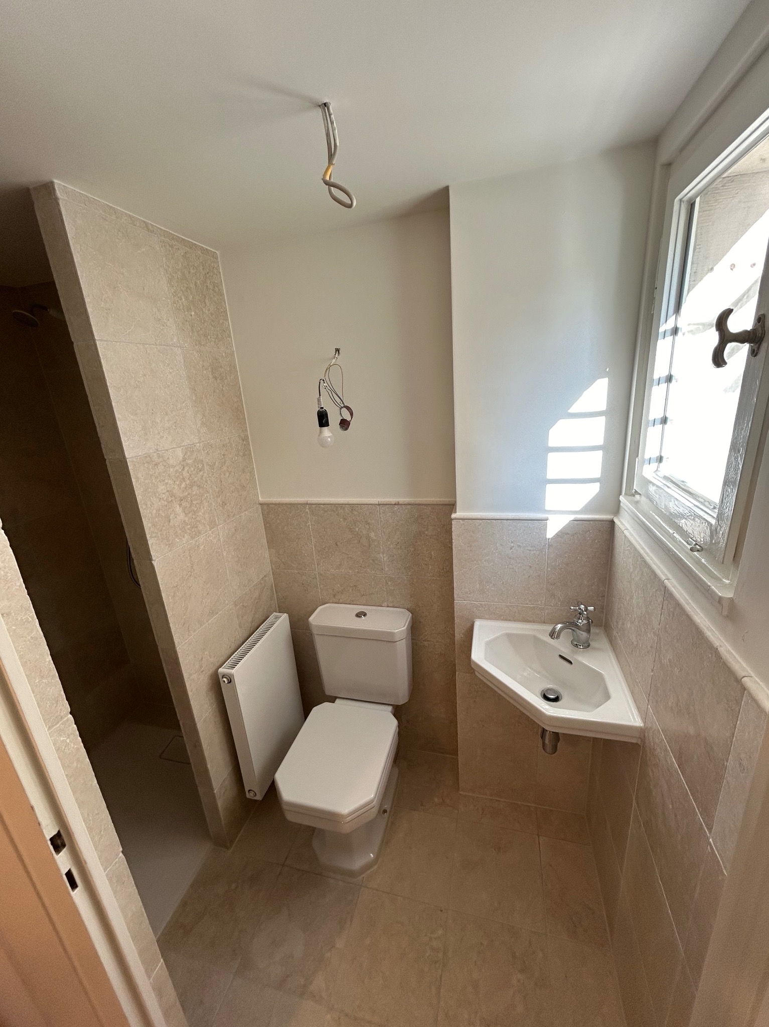 Jasna łazienka w trakcie prac wykończeniowych: beżowe płytki, biała armatura, widoczne kable. Okno z promieniami słońca. Ujęcie z perspektywy wejścia.