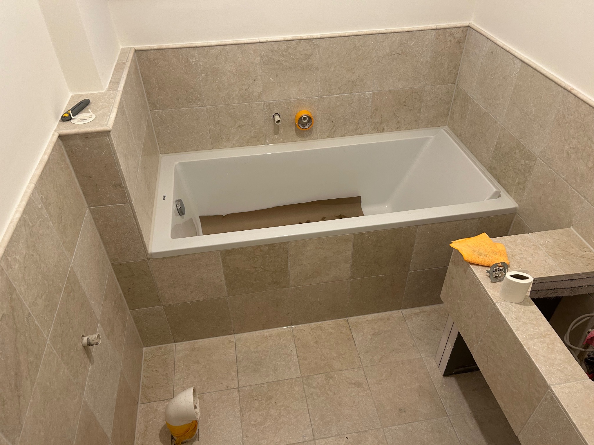 Jasna łazienka w trakcie remontu: beżowe płytki, biała wanna, widoczne narzędzia i akcesoria, etap prac wykończeniowych. Ujęcie z góry.