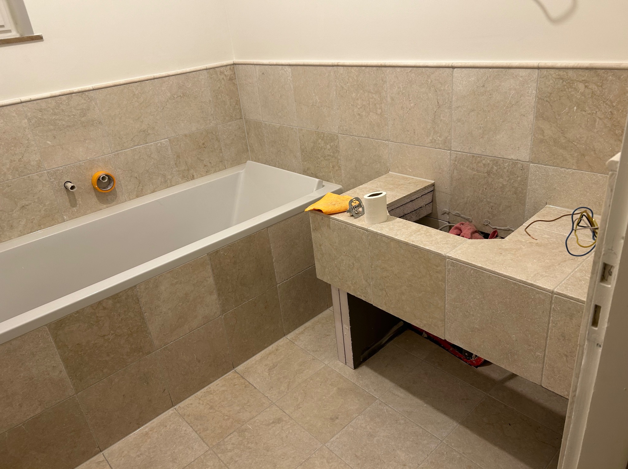 Beżowa łazienka w trakcie wykańczania: wanna, zabudowa umywalki z płytkami, widoczne instalacje. Na blacie leżą akcesoria. Neutralne oświetlenie.