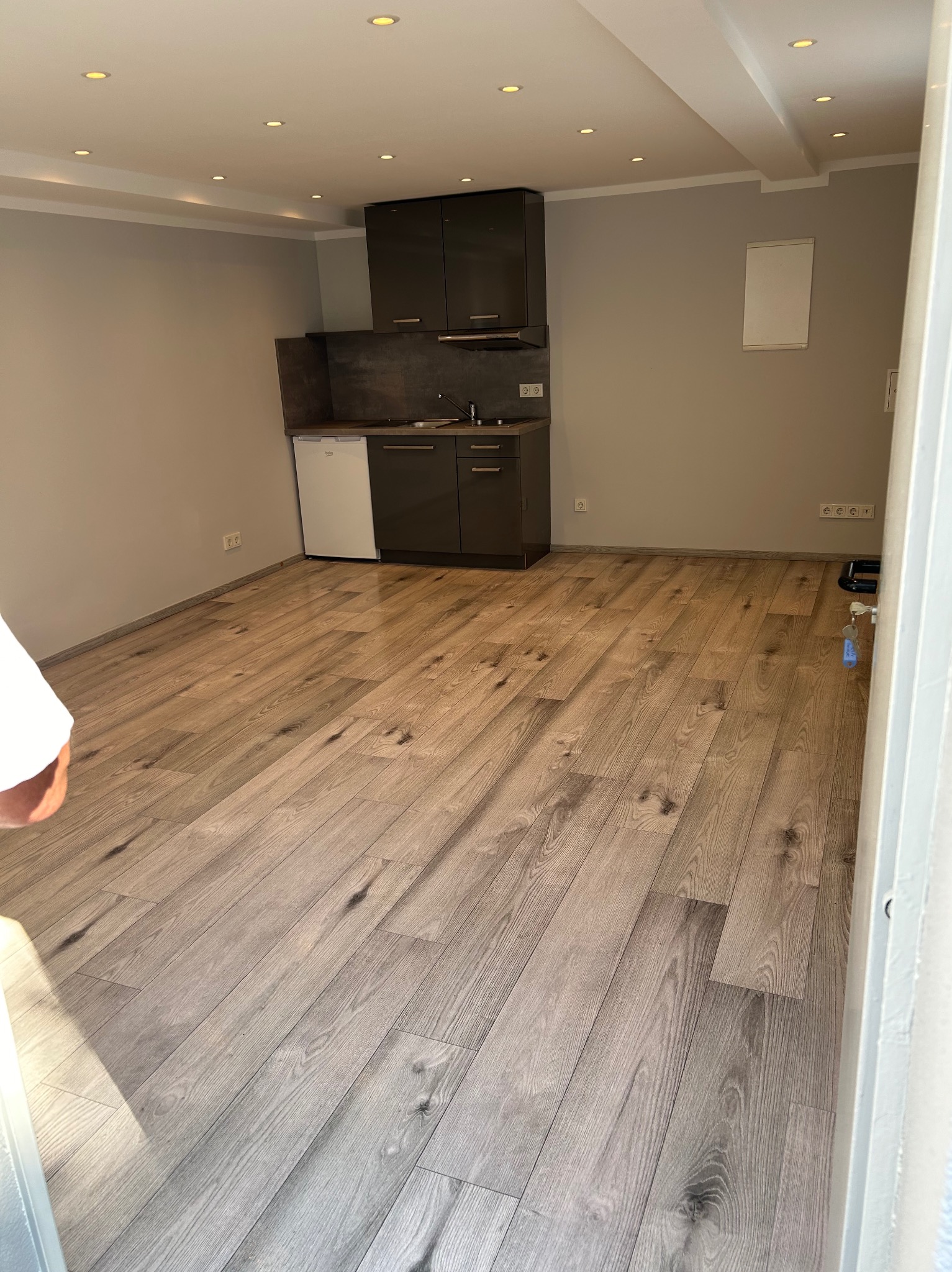 Jasne panele podłogowe w wyremontowanej kuchni z zabudową w kolorze grafitu i białym sprzętem AGD. Widok z perspektywy wejścia do pomieszczenia, oświetlenie punktowe.