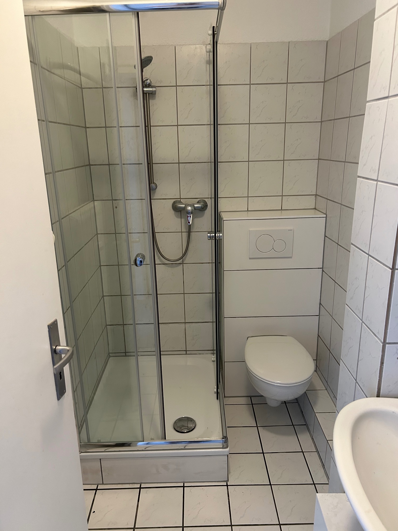 Biała łazienka z kabiną prysznicową, toaletą i umywalką. Widoczne białe płytki na ścianach i podłodze. Minimalistyczny styl, standardowe wyposażenie.