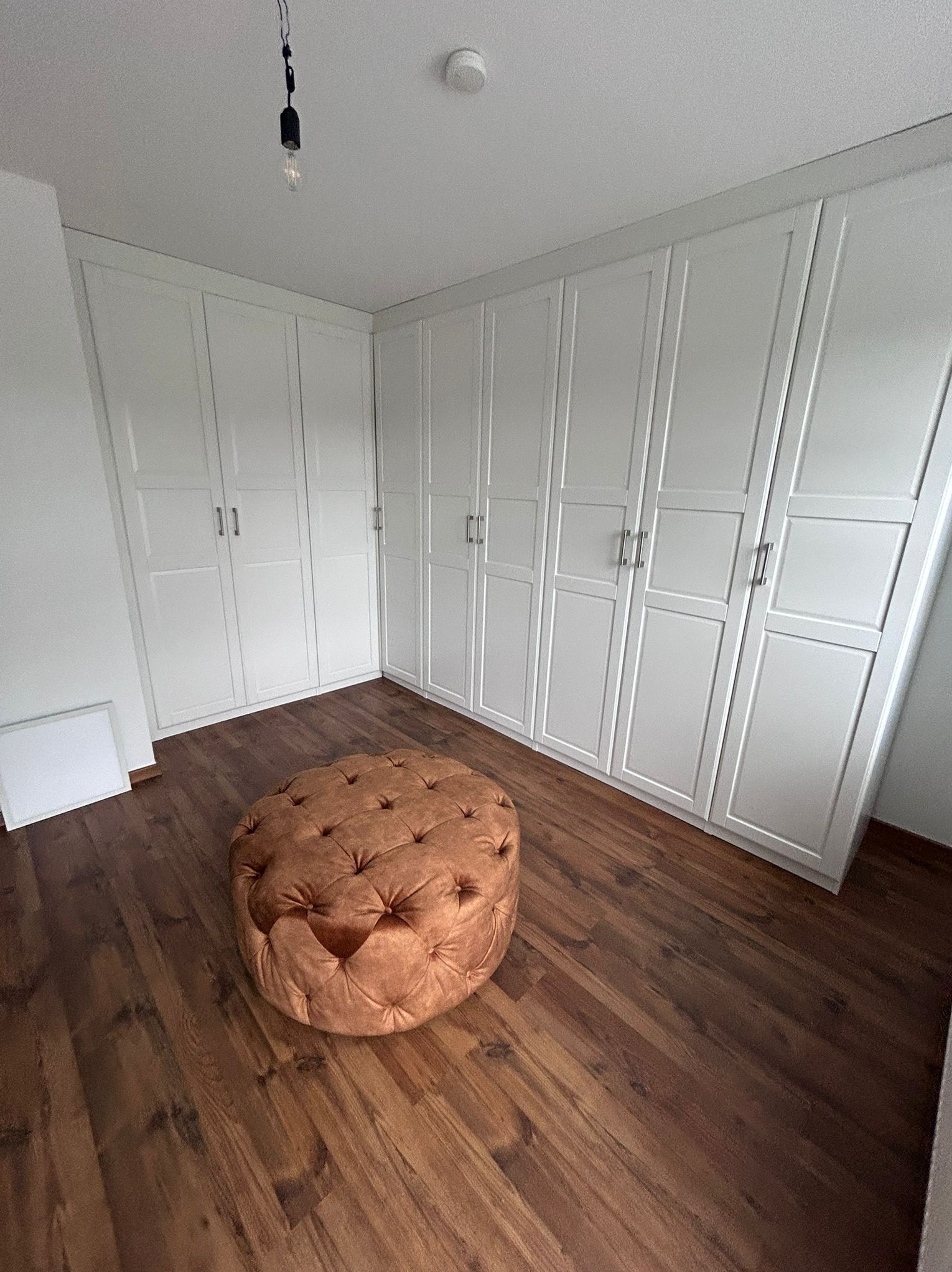 Białe szafy wnękowe z panelowymi drzwiami i brązowy puf na drewnianej podłodze. Minimalistyczne wnętrze z wiszącą żarówką.