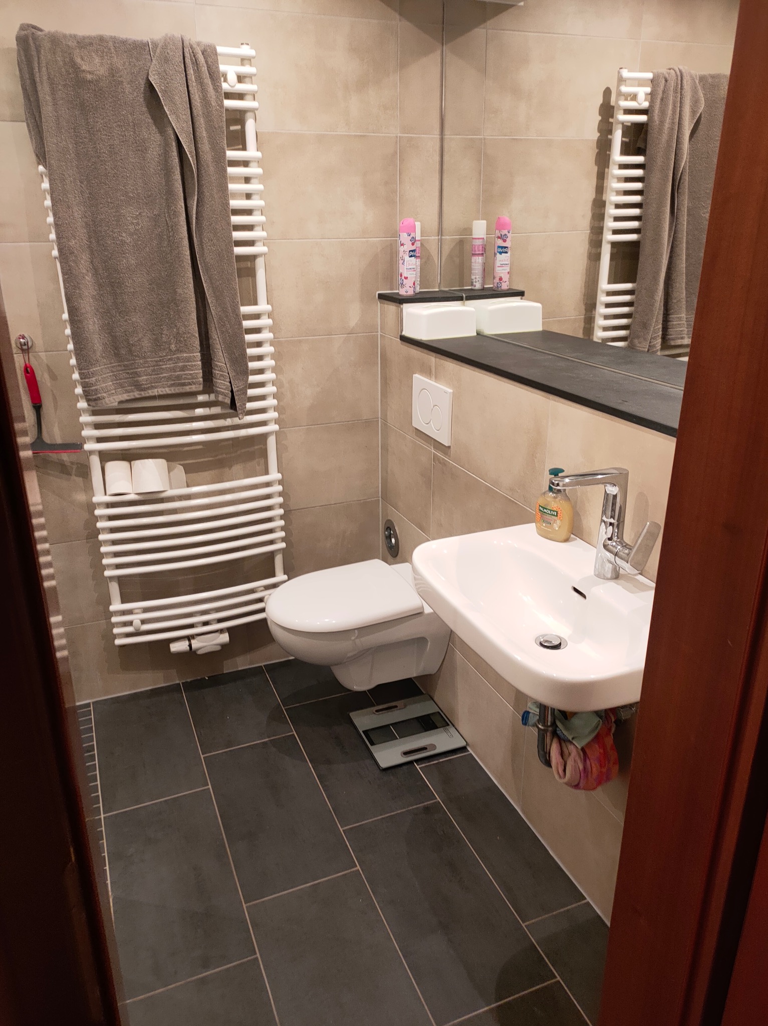 Jasna łazienka z beżowymi płytkami, grzejnikiem drabinkowym z ręcznikiem i umywalką. Na podłodze ciemne płytki. Uwagę przykuwa wisząca umywalka z baterią.