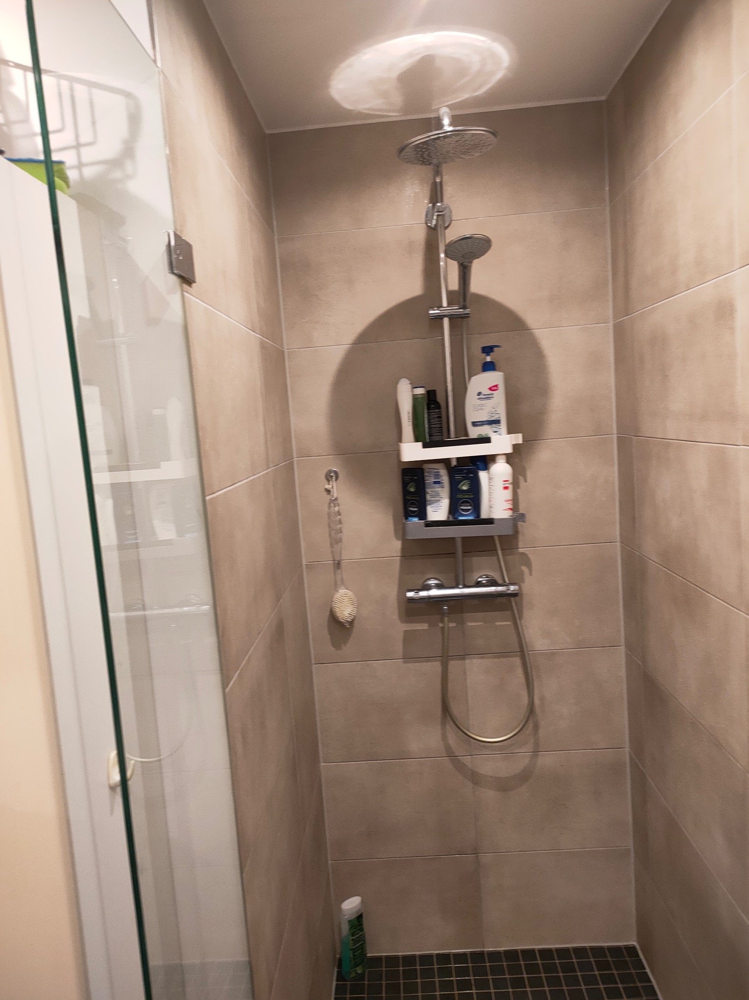 Kabina prysznicowa z deszczownicą i półką na kosmetyki, wykończona beżowymi płytkami. Widoczny fragment czarnej mozaiki podłogowej i akcesoria kąpielowe.