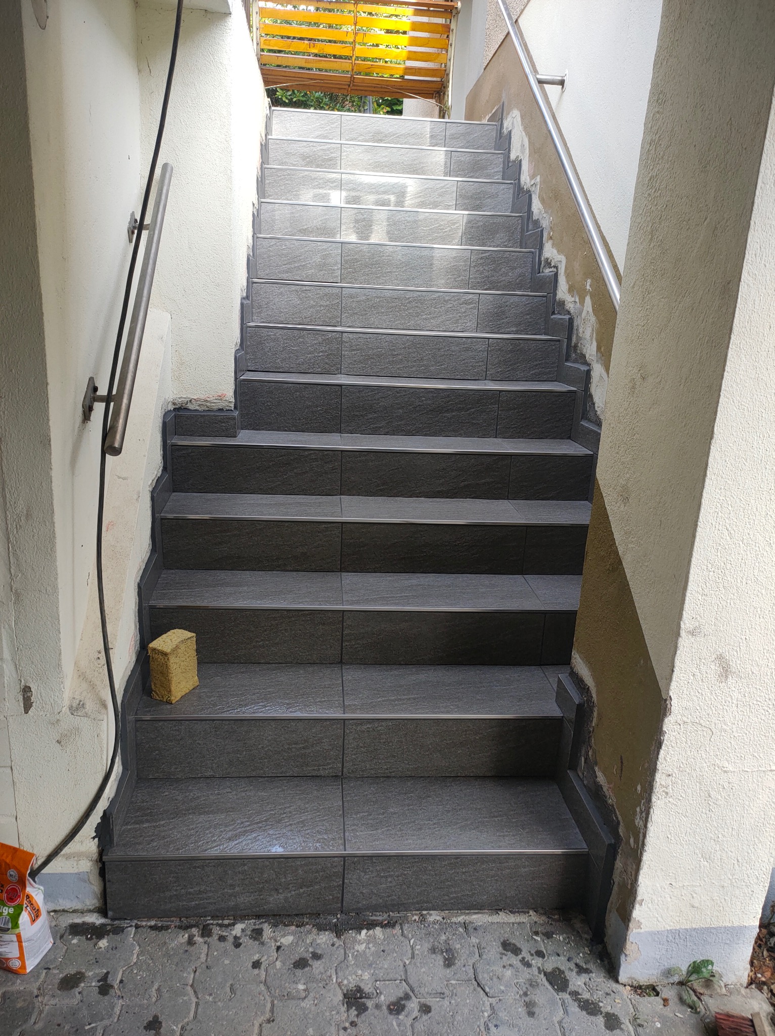 Wykończone schody zewnętrzne z szarych płytek, z metalowymi listwami, prowadzące do góry. Widoczna gąbka glazurnicza na schodach i ślady po pracach wykończeniowych na ścianach.