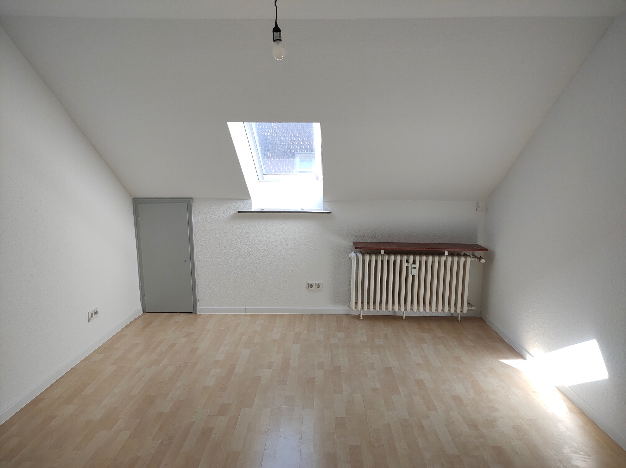 Puste, białe poddasze z drewnianą podłogą, oknem dachowym, grzejnikiem i szarą ścianą. Widoczne gniazdka elektryczne i żarówka na kablu.