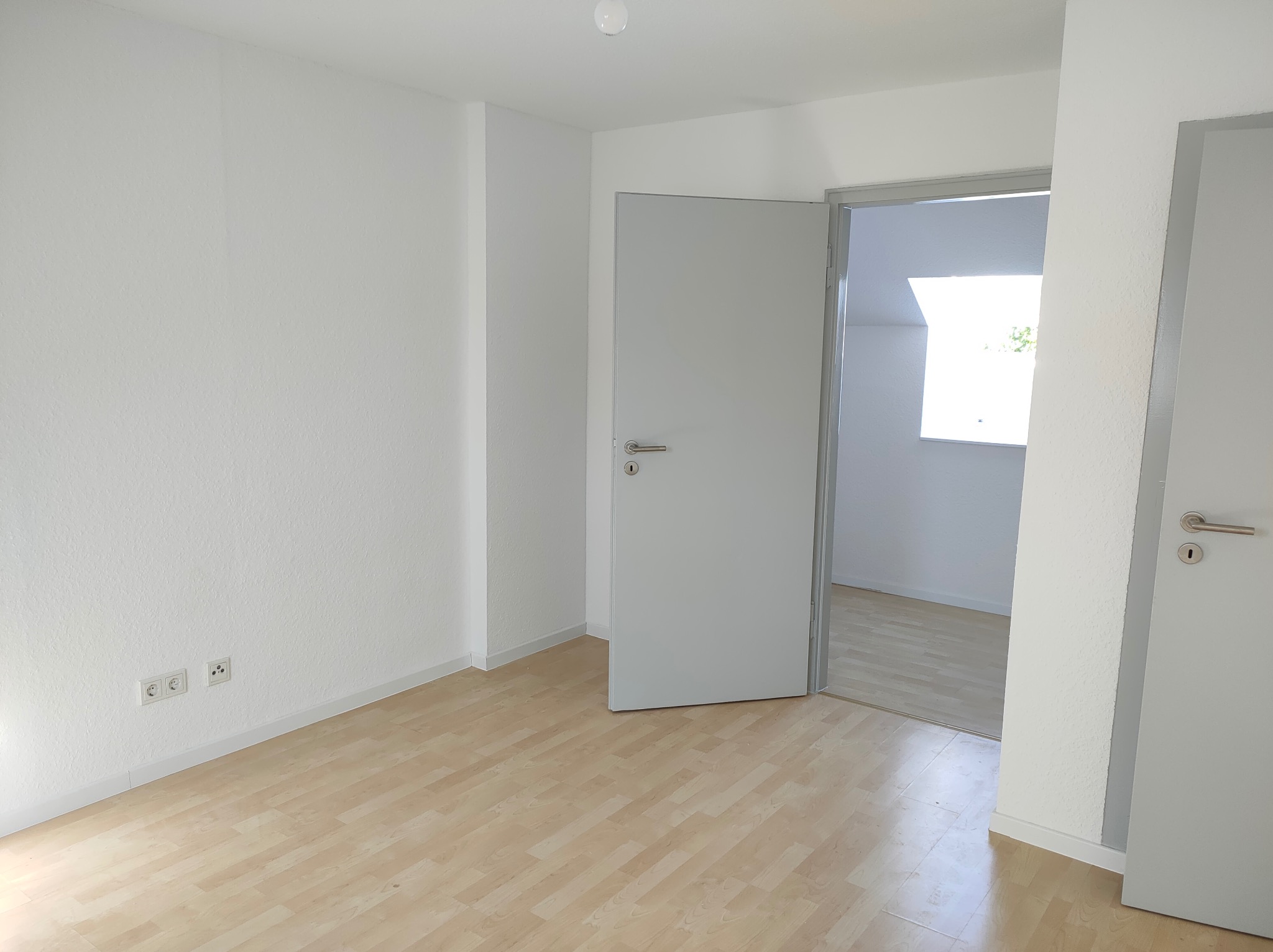 Jasne wnętrze z parkietem i białymi ścianami. Otwarta, szara  płycinowa  furtka zaprasza do kolejnego pomieszczenia. Widoczne gniazdka elektryczne.