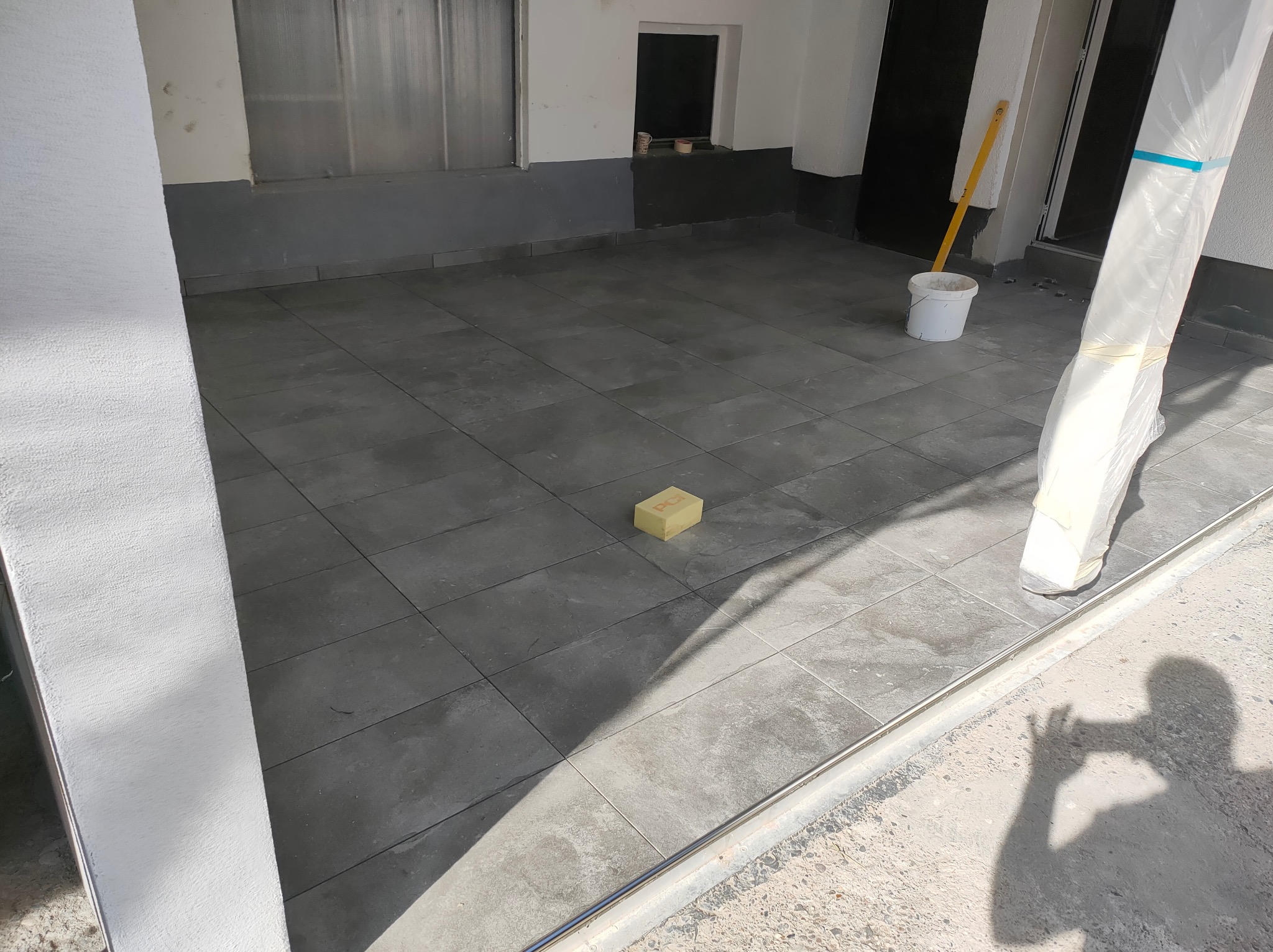 Ułożona, szara terakota w garażu lub na balkonie. Widoczny poziomica oparta o ścianę, wiadro i cień osoby robiącej zdjęcie. Kostka do czyszczenia fug na podłodze.