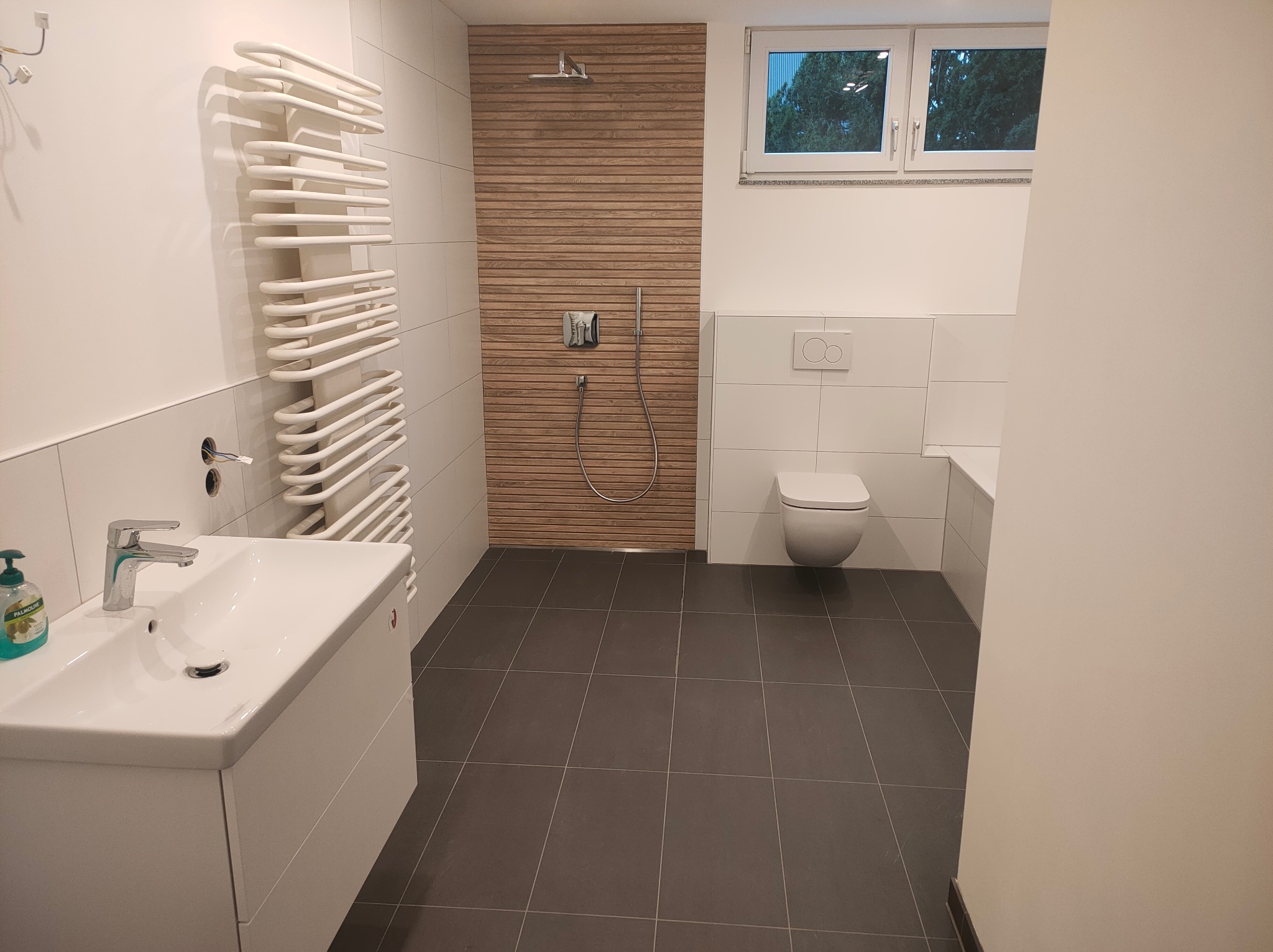 Nowoczesna łazienka z drewnianym panelem prysznicowym, grzejnikiem drabinkowym i szarą podłogą. Minimalistyczny design, biała armatura i okno z widokiem na zieleń.