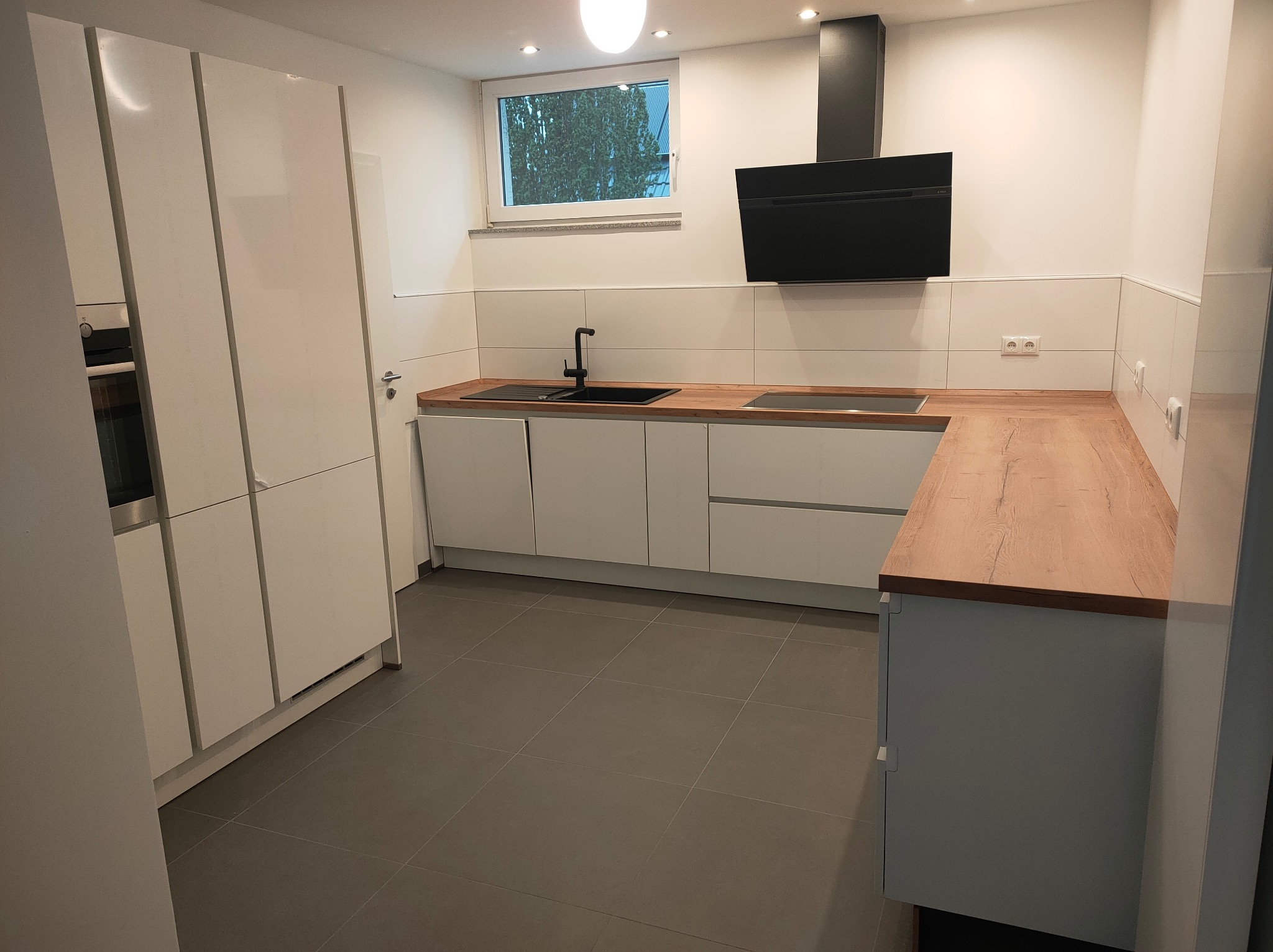 Nowoczesna, jasna kuchnia z białymi szafkami, drewnianym blatem i czarną armaturą. Minimalistyczny design z płytkami na ścianie i szarą podłogą.