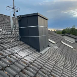 Czarny komin wentylacyjny na szarym dachu z dachówki ceramicznej, zabezpieczony metalowymi obejmami, antena TV w tle, pochmurne niebo.