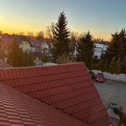 MajsterDACH - Czerwona dachówka karpiówka w perspektywie z góry, częściowo umyta przez osobę w czarnym ubraniu. W tle zabudowa miejska i zachodzące słońce.