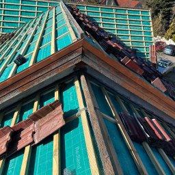MajsterDACH - Fragment dachu w trakcie prac. Widoczna drewniana konstrukcja, membrana w kolorze turkusowym i częściowo ułożona dachówka w odcieniach czerwieni i brązu. Prace dekarskie w toku.