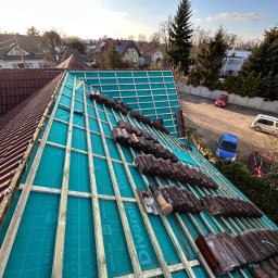 MajsterDACH - Układanie dachówki na łatach na folii paroizolacyjnej z widocznymi napisami producenta. Częściowo pokryty dach z perspektywy z góry, w tle zabudowa jednorodzinna.