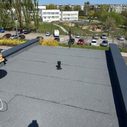 MAJSTERDACH VOLODYMYR HUMENIUK - Szary, płaski dach pokryty papą termozgrzewalną z kominkiem wentylacyjnym na tle zabudowy miejskiej i błękitnego nieba w słoneczny dzień. Widoczny cień osoby robiącej zdjęcie.
