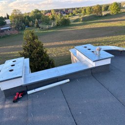 MAJSTERDACH VOLODYMYR HUMENIUK - Szary dach z papy z kominami obłożonymi blachą, na dachu leżą czerwone rękawice robocze i zwinięta siatka. W tle zieleń drzew i łąk w słoneczny dzień.