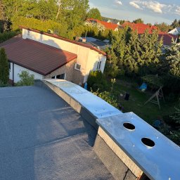 MAJSTERDACH VOLODYMYR HUMENIUK - Szary dach z blachy z dwoma otworami wentylacyjnymi, widok z góry na tle zieleni i zabudowań. Metalowe wykończenie komina. Słoneczny dzień.