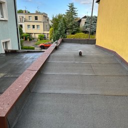 MAJSTERDACH VOLODYMYR HUMENIUK - Płaski dach pokryty ciemną papą termozgrzewalną z widocznymi łączeniami i kominkiem wentylacyjnym. Brązowe listwy krawędziowe z kroplami deszczu, w tle osiedle domów.
