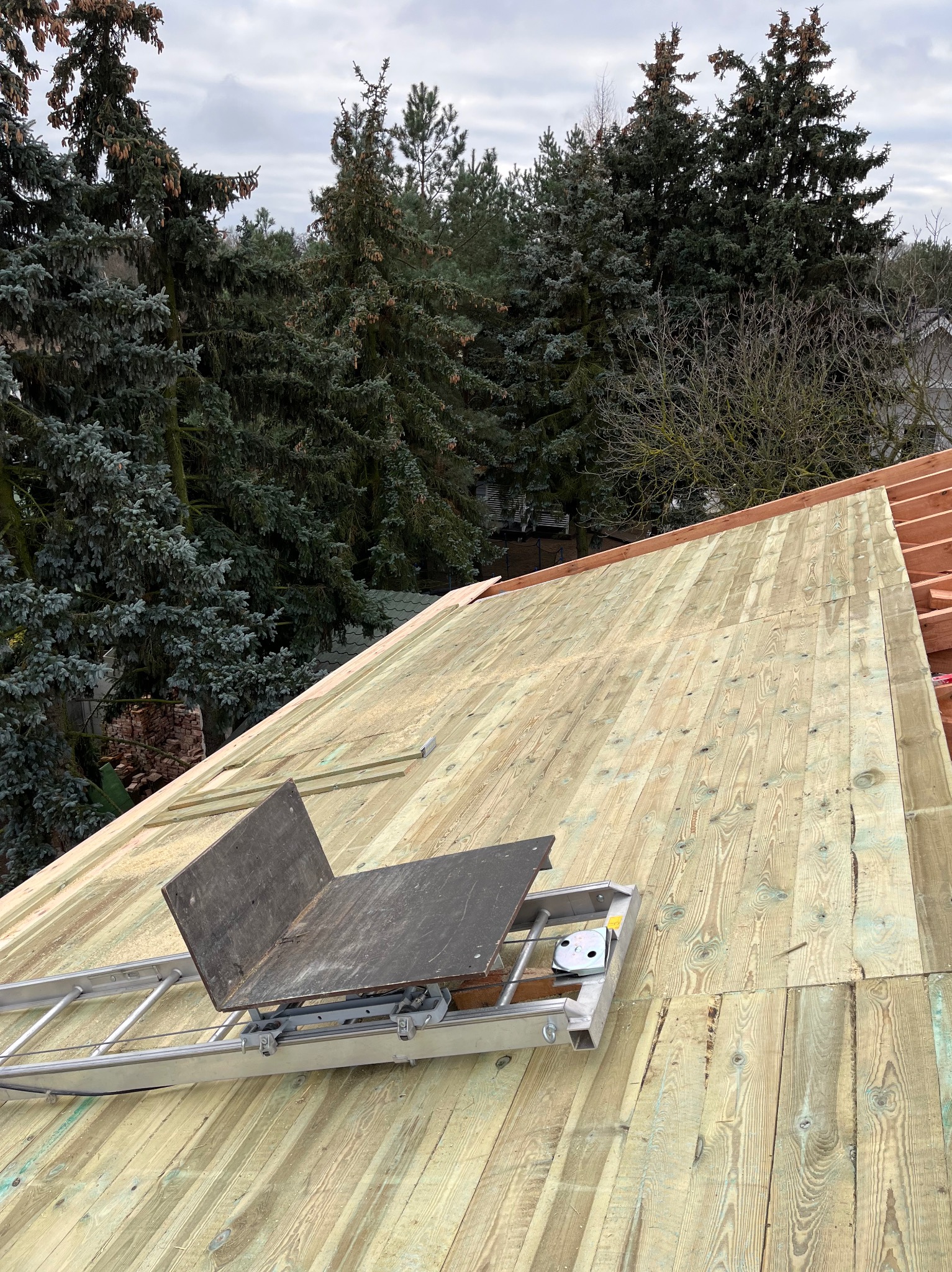 Drewniane poszycie dachu z widoczną konstrukcją więźby i specjalistycznym podestem na drabinie, tle drzewa iglaste i pochmurne niebo.