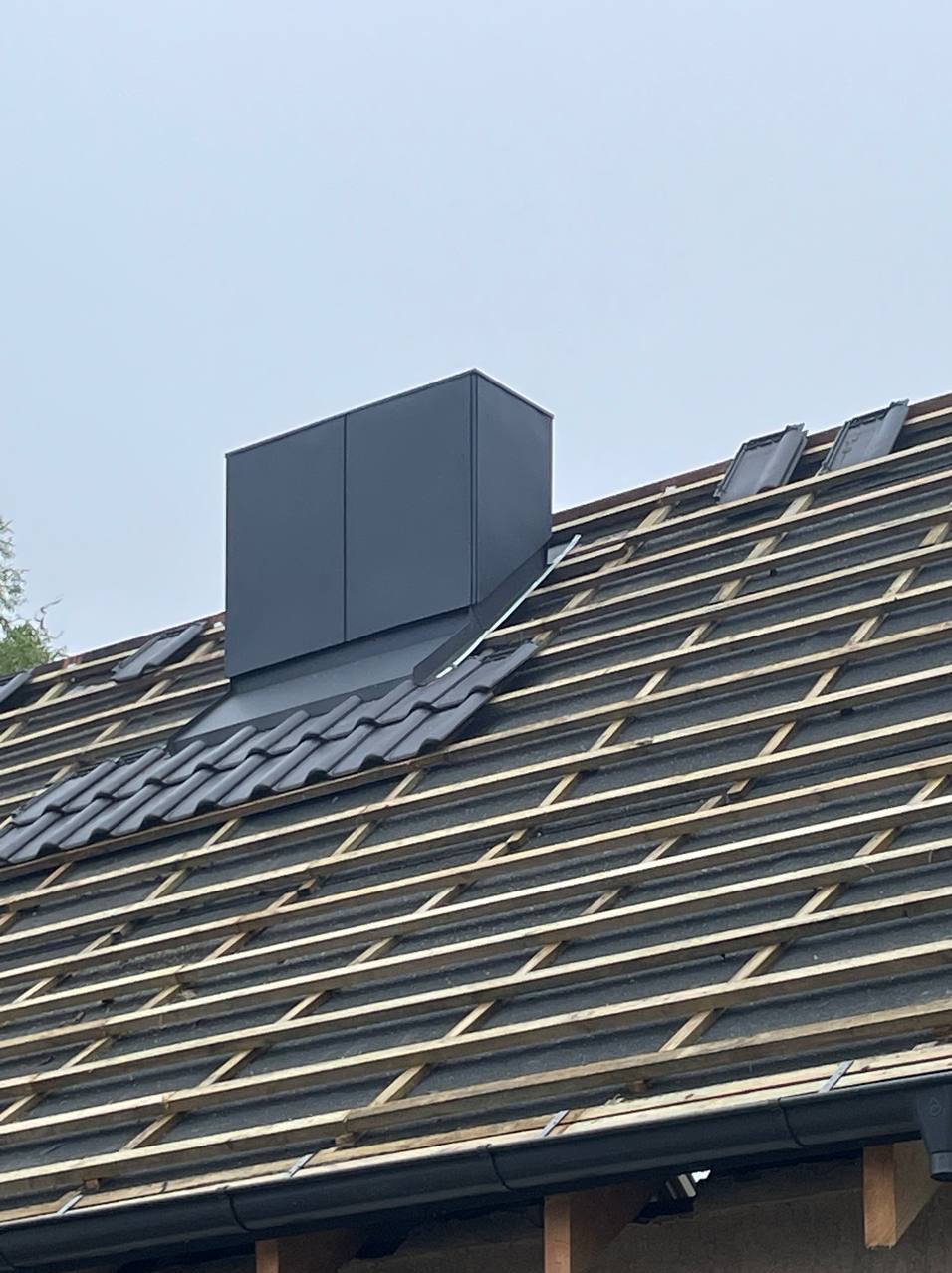 Fragment dachu z zamontowanym, ciemnym kominem wentylacyjnym. Widoczna drewniana konstrukcja, częściowo pokryta dachówką ceramiczną w kolorze antracytowym.