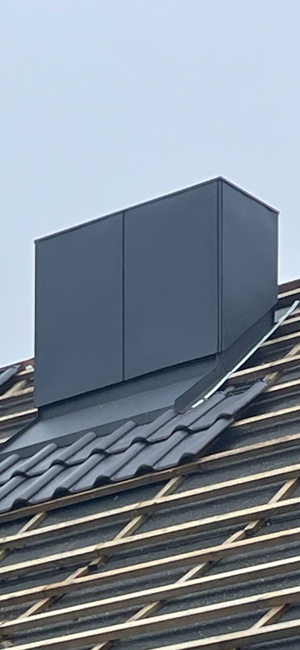 Szary komin wentylacyjny na dachu z ciemnej dachówki ceramicznej, widoczne łaty i kontrłaty, pochmurne niebo w tle.