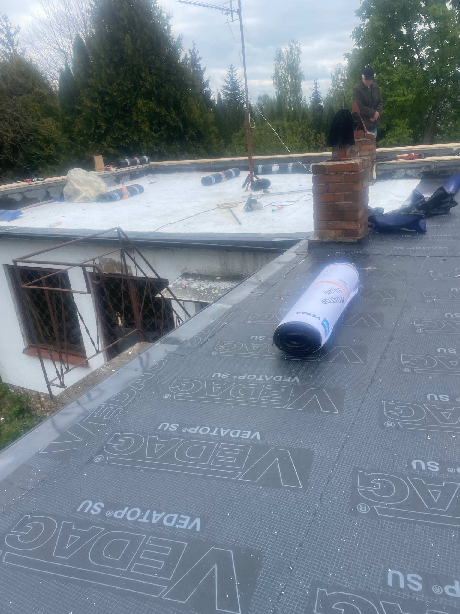 Płaski dach w trakcie krycia papą termozgrzewalną Vedag, widoczny komin z cegły i pracownik w oddali. W tle zieleń drzew i krzewów.