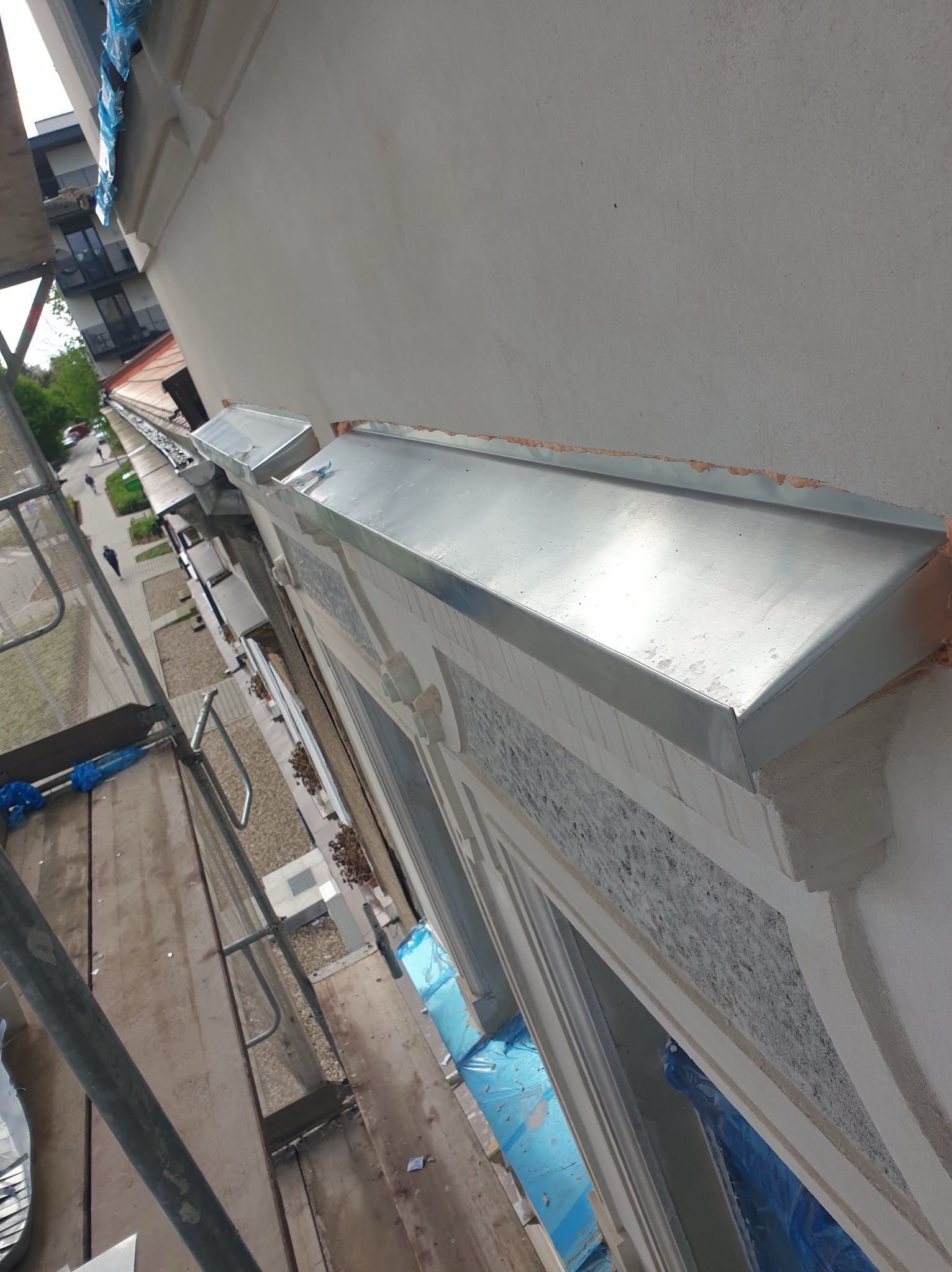 Szczegółowe ujęcie z rusztowania na nową, błyszczącą obróbkę blacharską okapową na budynku z dekoracyjnymi elementami architektonicznymi.