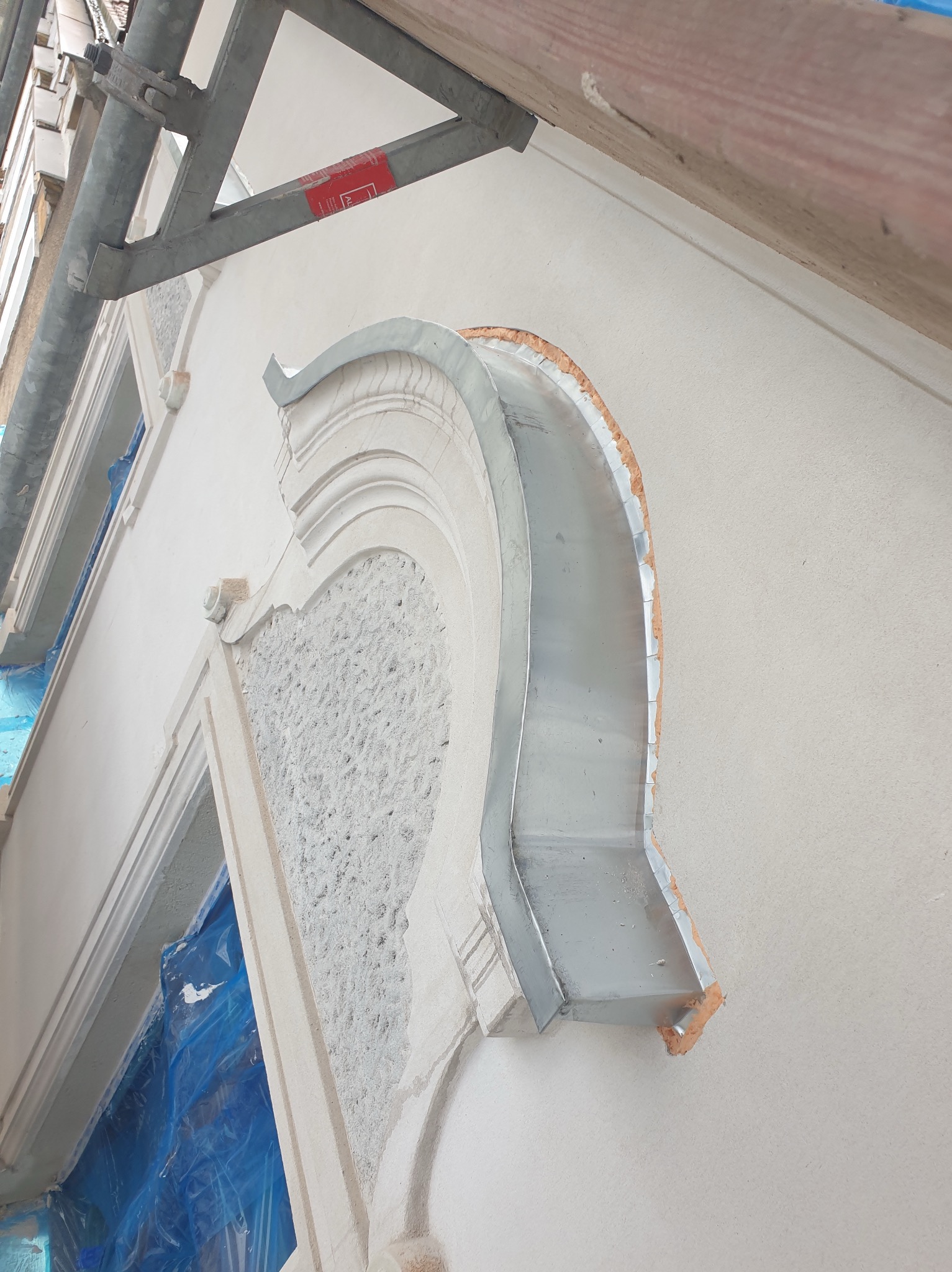 Detal elewacji budynku w trakcie renowacji: dekoracyjny element architektoniczny z blachy, zamontowany na ścianie pokrytej jasnym tynkiem. Widoczne rusztowanie.