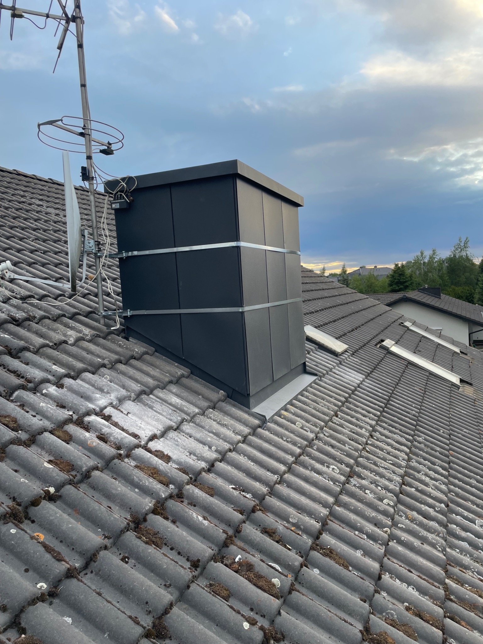 Czarny komin wentylacyjny na szarym dachu z dachówki ceramicznej, zabezpieczony metalowymi obejmami, antena TV w tle, pochmurne niebo.