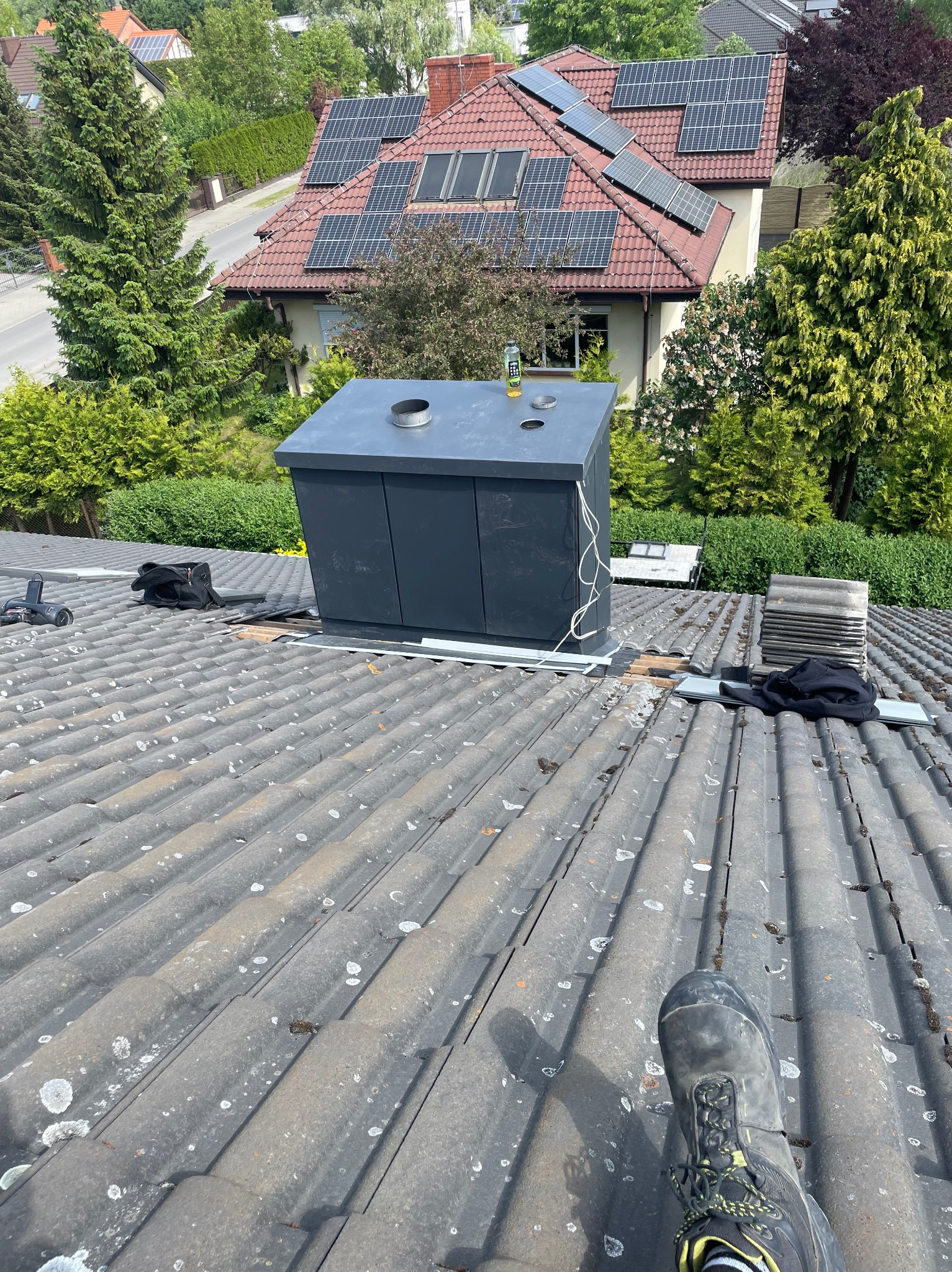 Montaż obudowy komina na dachu z szarej dachówki. Widoczny fragment buta montera, narzędzia, budynek z panelami słonecznymi w tle, zieleń.