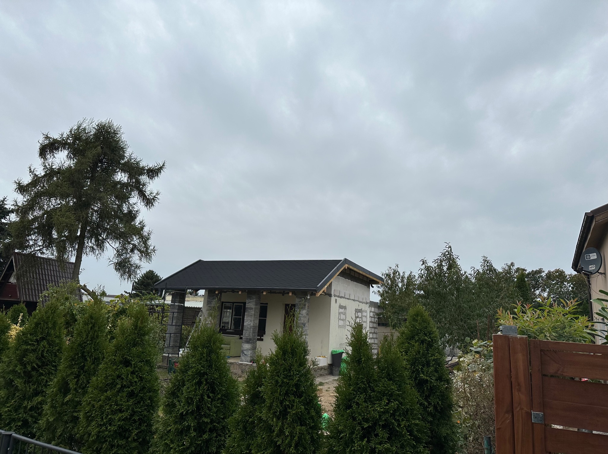 Dom z czarnym dachem w trakcie budowy, widoczny zza rzędu iglaków. Część ścian jest otynkowana, część w surowym stanie. Pochmurne niebo w tle.