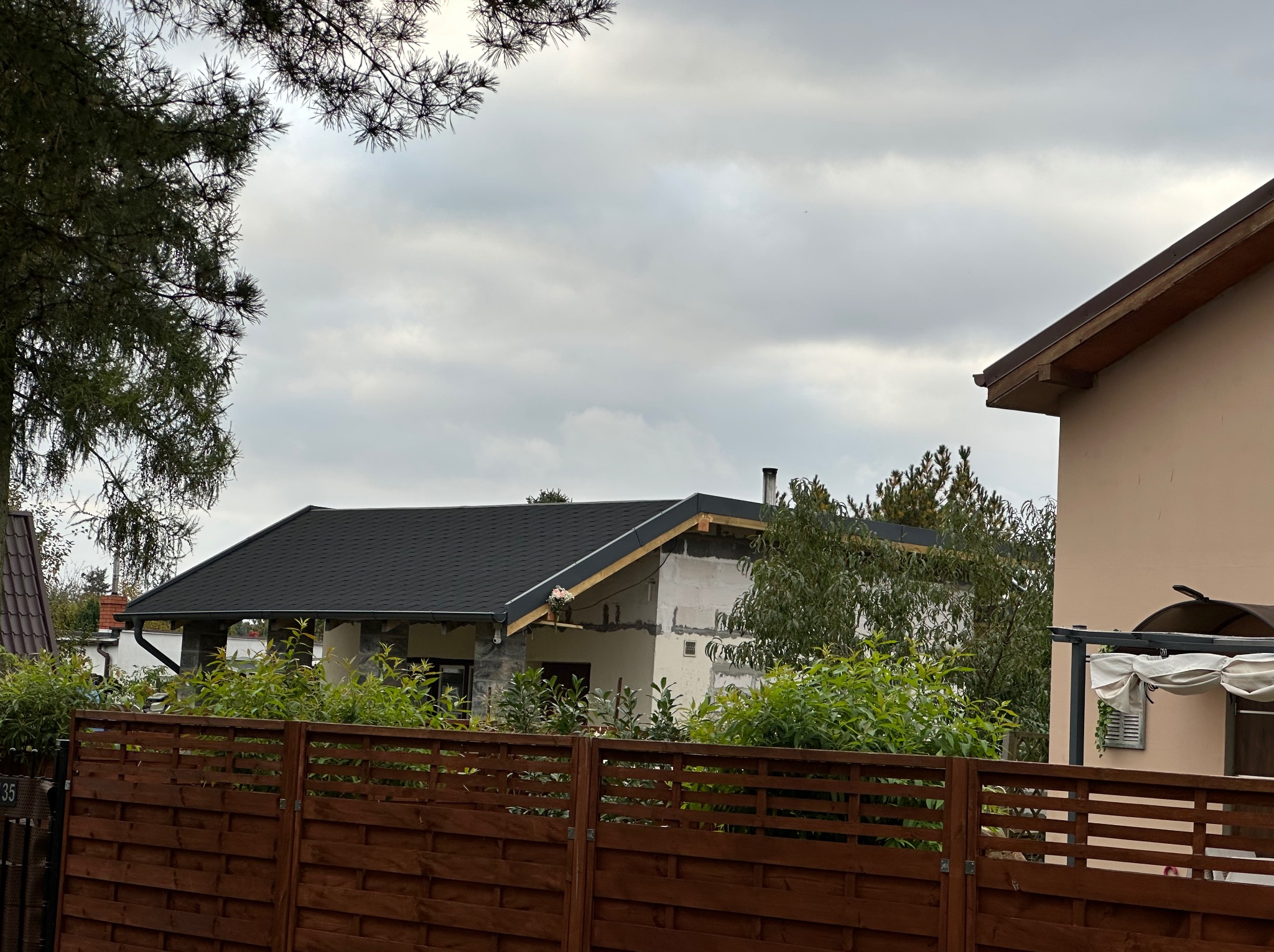 Dom w trakcie budowy z nowym, czarnym dachem, widoczny zza drewnianego ogrodzenia. Fragment innego budynku po prawej, pochmurne niebo.