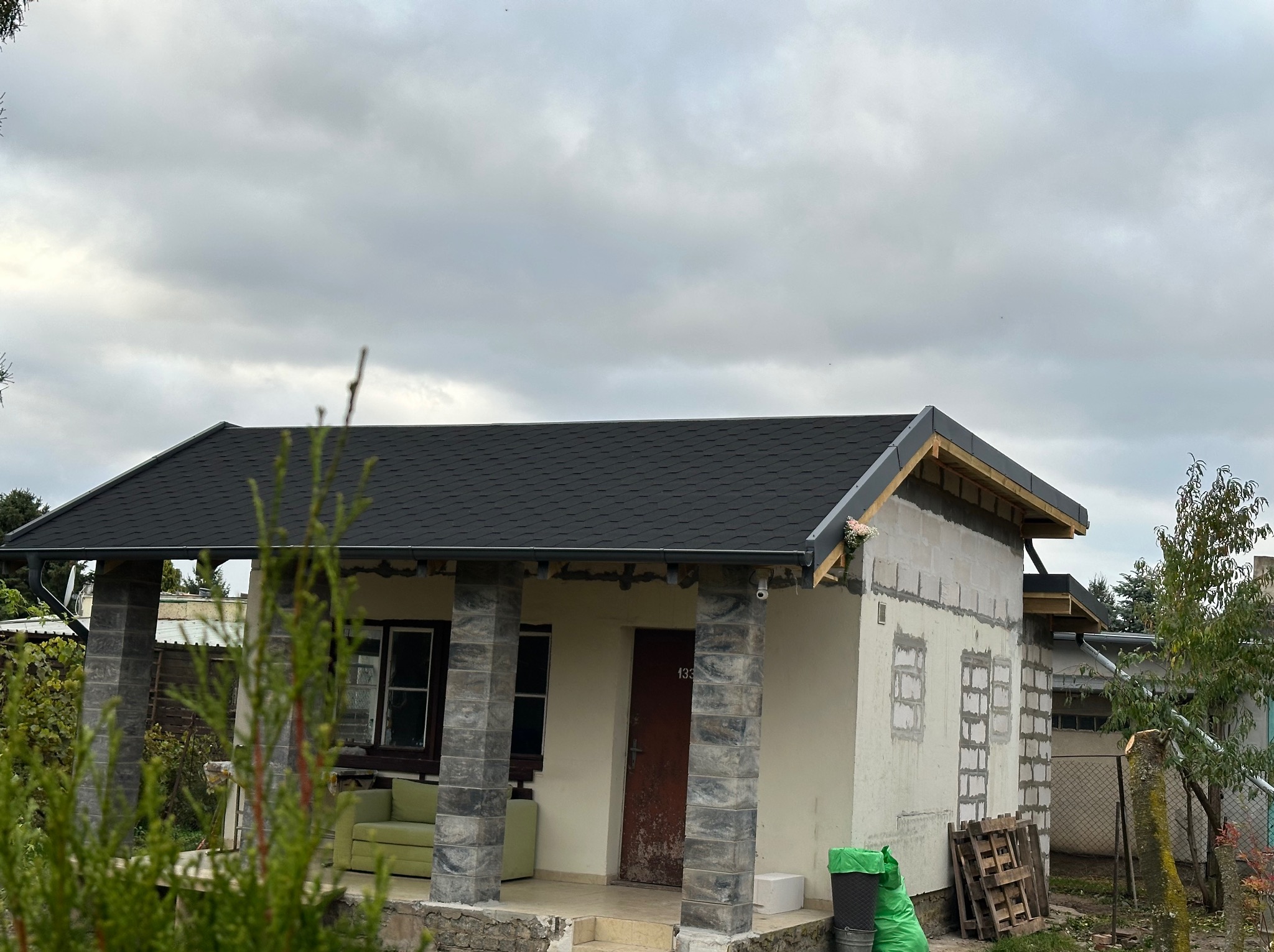 Dom w trakcie budowy z nowym, czarnym dachem z papy bitumicznej. Widoczne rynny i fragment elewacji w surowym stanie.