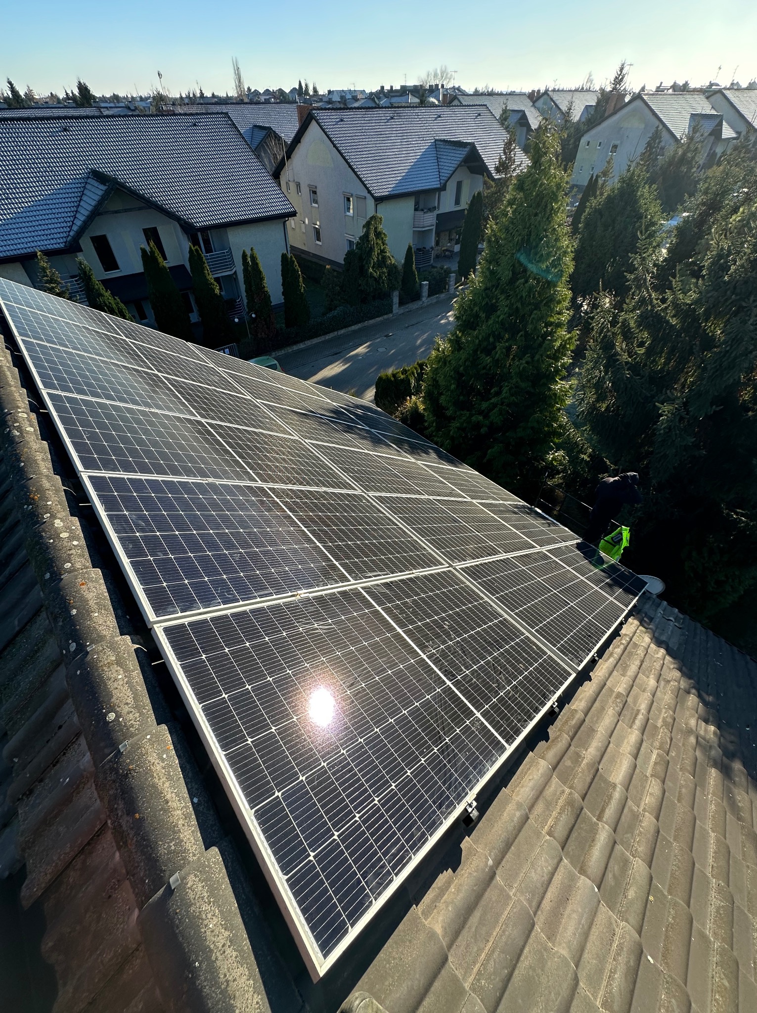 Panel słoneczny na dachu domu jednorodzinnego w słoneczny dzień. Widoczne dachówki i okoliczne budynki. Instalacja fotowoltaiczna na ukośnym dachu.
