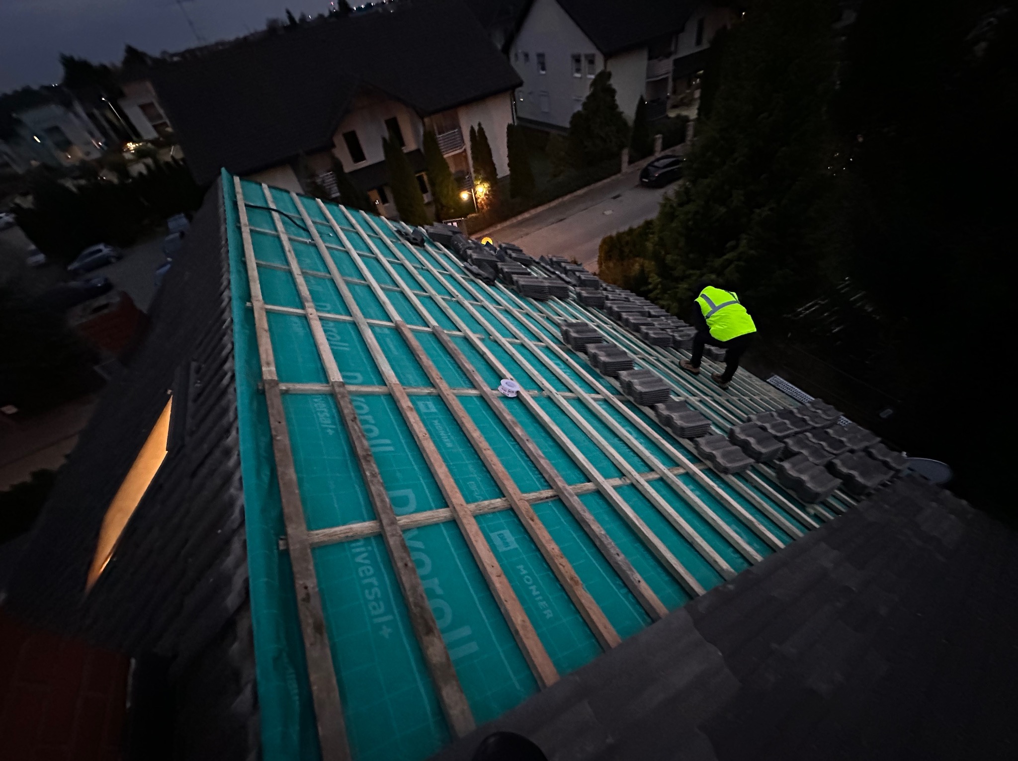 Układanie dachówki na przygotowanej konstrukcji dachu z membraną, widoczny dekarz w odblaskowej kamizelce. Prace dekarskie w trakcie, wieczorne oświetlenie.