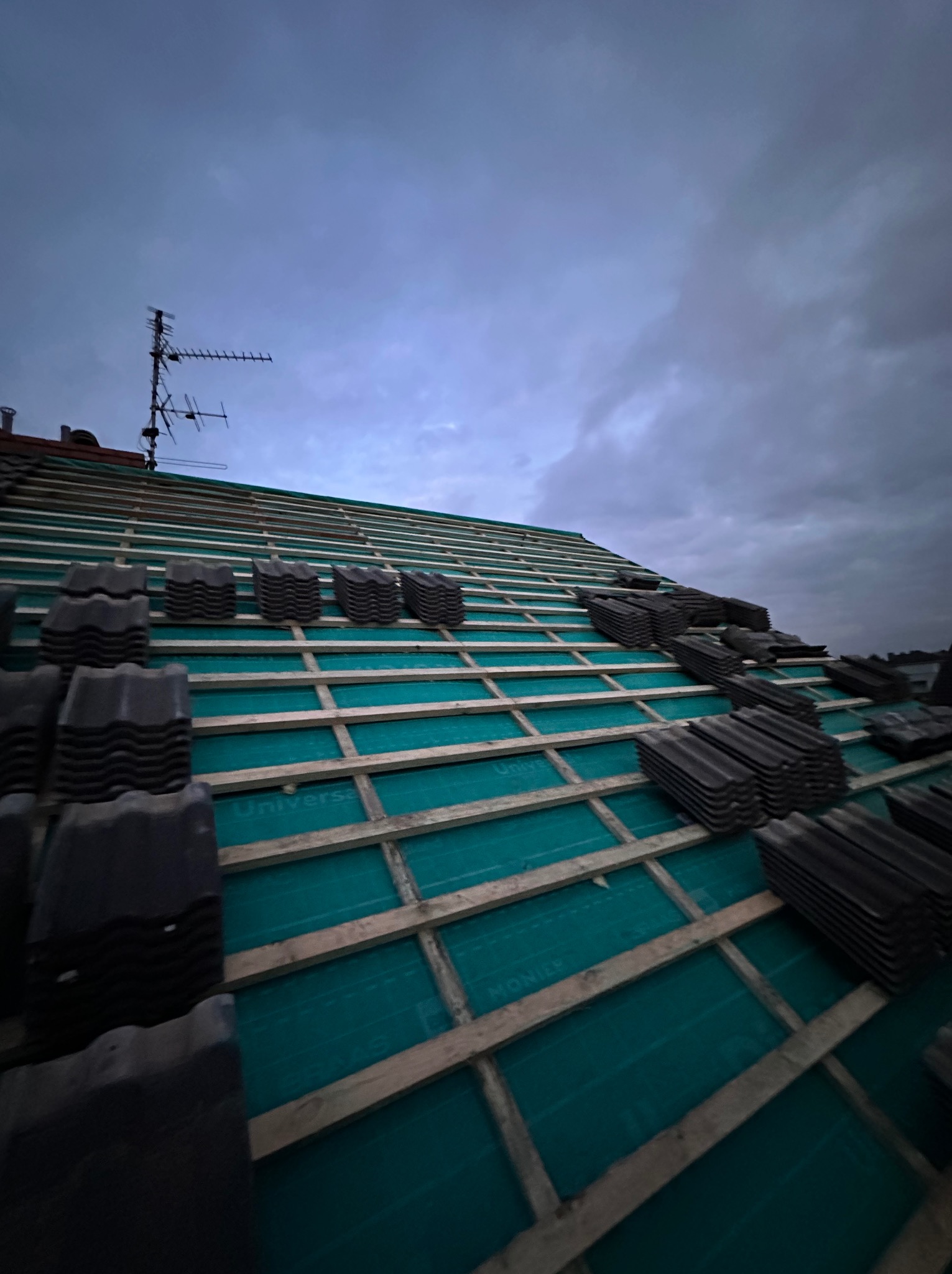 Układanie dachówki na zielonej membranie na drewnianej konstrukcji dachu w pochmurny dzień. Widoczna antena na dachu sąsiedniego budynku.