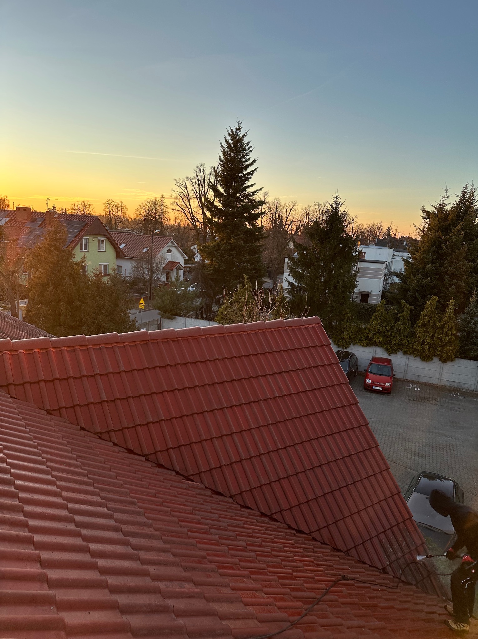 Czerwona dachówka karpiówka w perspektywie z góry, częściowo umyta przez osobę w czarnym ubraniu. W tle zabudowa miejska i zachodzące słońce.