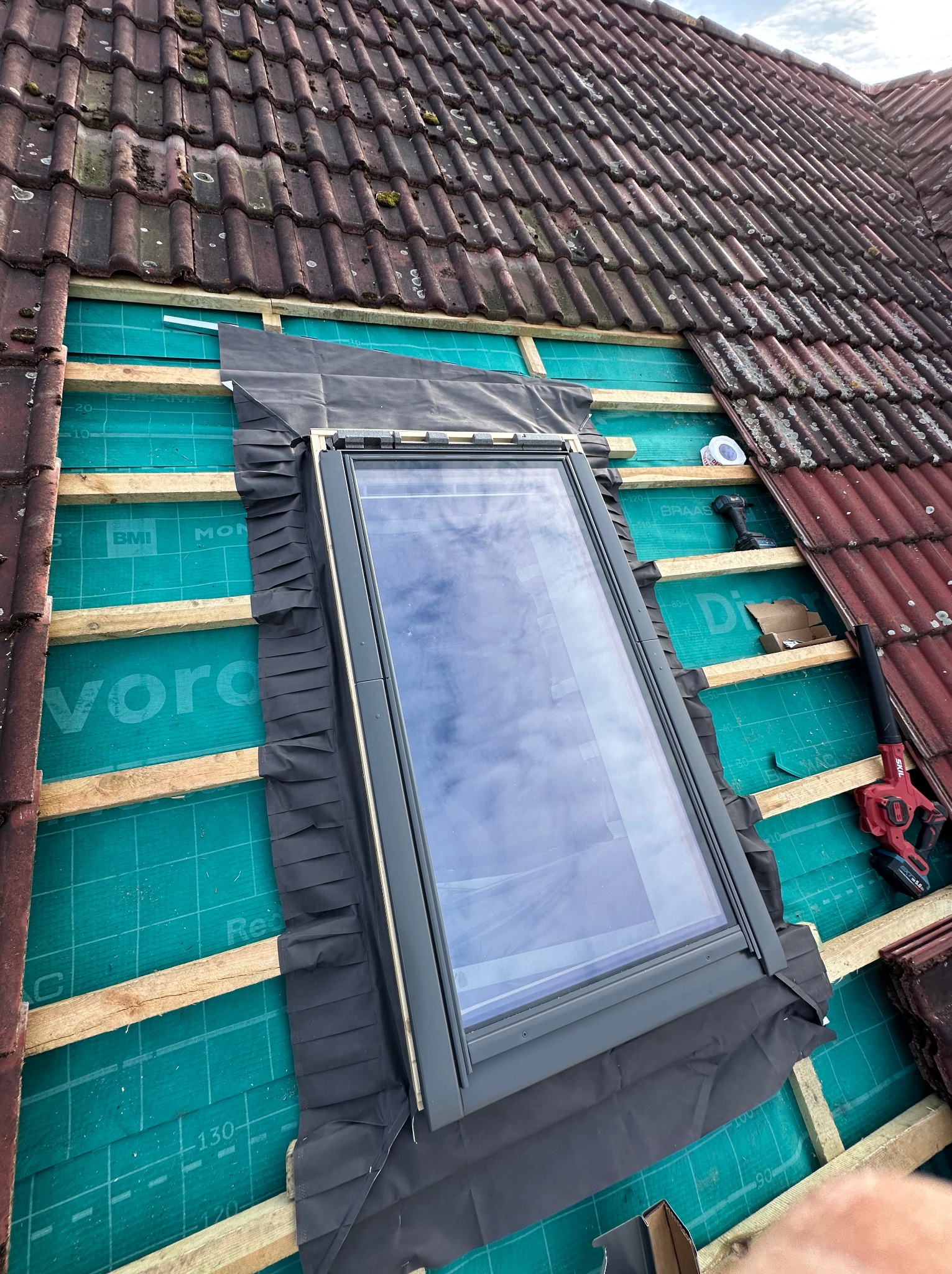 Montaż okna dachowego na zielonej membranie z widocznymi łatami i częścią starego, brązowego pokrycia. Narzędzia pracy w tle. Ujęcie z góry.